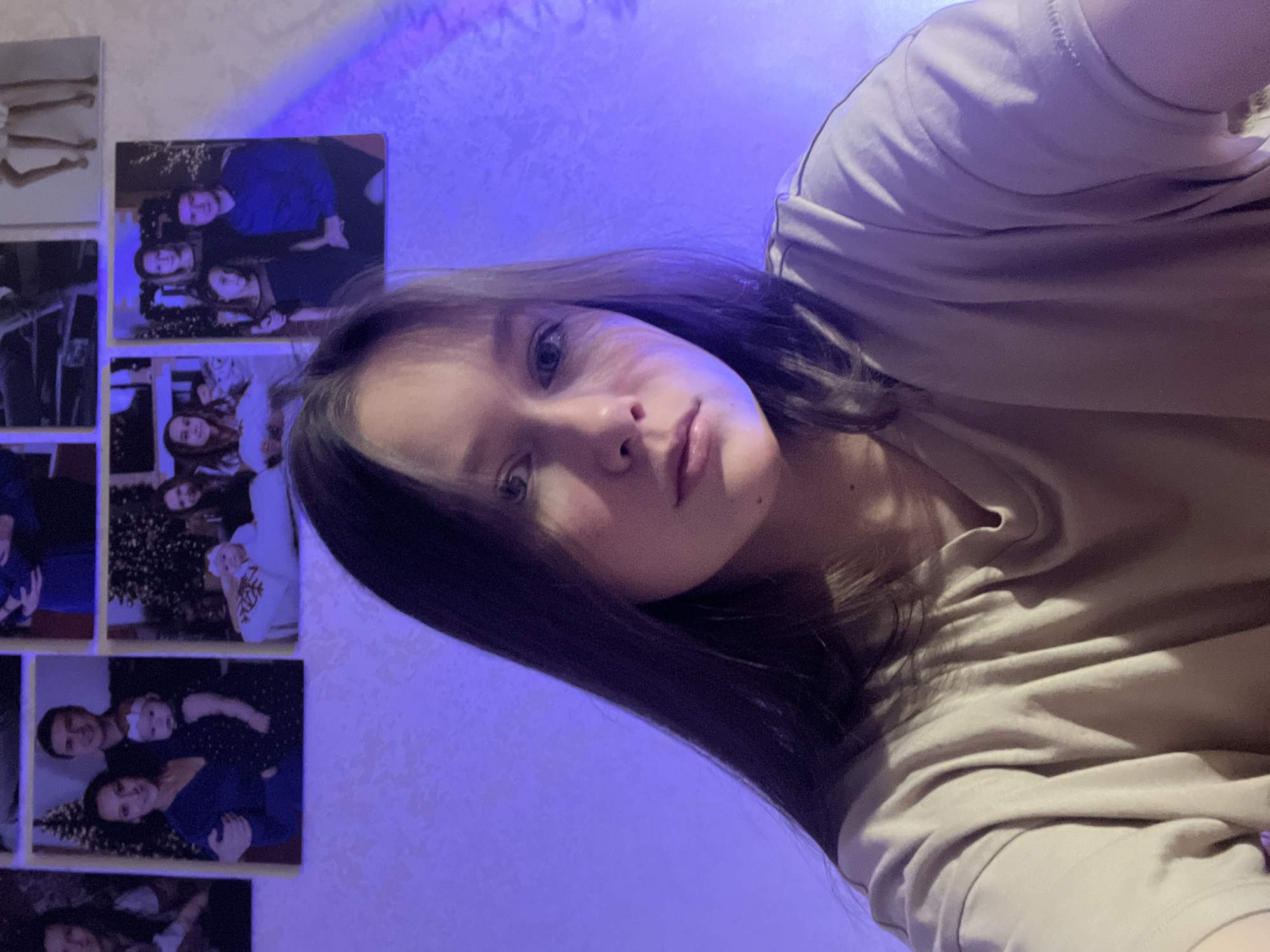 A young woman is pictured indoors in Chaikovsky, Russia. The photo is taken at an upward angle, showing her face, part of her torso in a light-colored t-shirt, and a wall behind her. The wall features a collage of printed photographs, including portraits of people, some with a Christmas tree. The room is lit with a blue light, creating a blue cast on the scene.