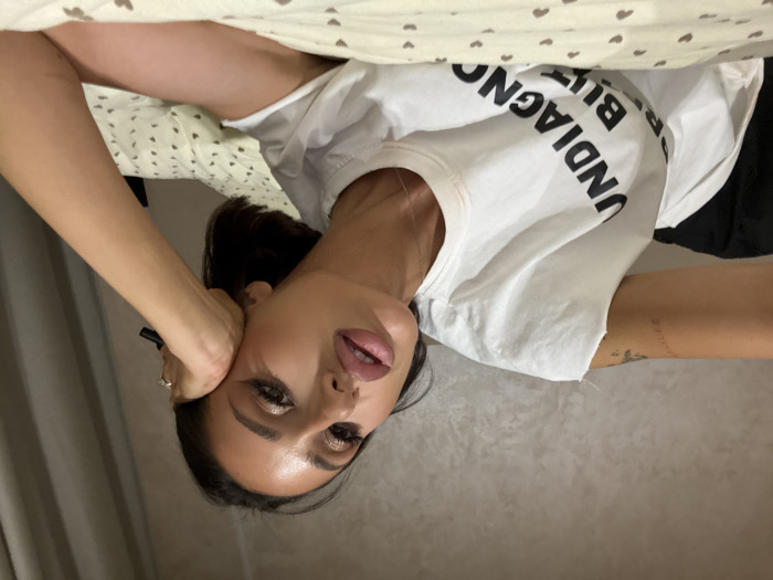 An adult female is depicted in a horizontal close-up, lying on a bed in an indoor residential setting in Khimki, Russia. She has short, dark hair and her face features prominent makeup, including winged eyeliner and plumped lips with a glossy finish. She wears a white t-shirt with a raw-edge left sleeve, displaying partially visible black text that reads "UNDIAGNOSED BUT". A tattoo is visible on her upper left arm. Her right hand, wearing a ring on the ring finger, supports her head, and she holds a slender dark object, possibly a pen, between her fingers. The bed is covered with light-colored bedding featuring a repeating pattern of small dark heart shapes. The background consists of a textured, light-toned wall and a darker curtain to the right.