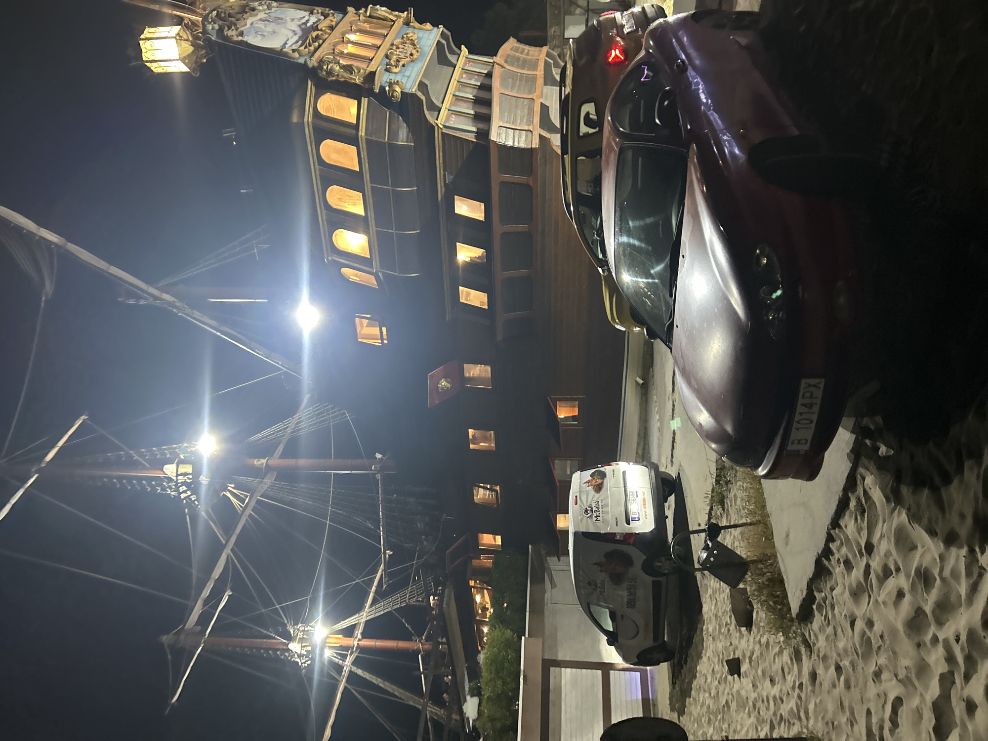 The image depicts a nighttime scene in Varna, Bulgaria, featuring a replica pirate ship, likely a restaurant or attraction, as the primary background. The ship is illuminated, with warm light emanating from its windows and decorative lanterns. In the foreground, three vehicles are parked on a sandy area. From left to right, the first vehicle is a white van with company logos and contact information visible on its rear. Next to it is a yellow SUV. The third vehicle, in the immediate foreground, is a red coupe. The ground is a mixture of sand and pavement.