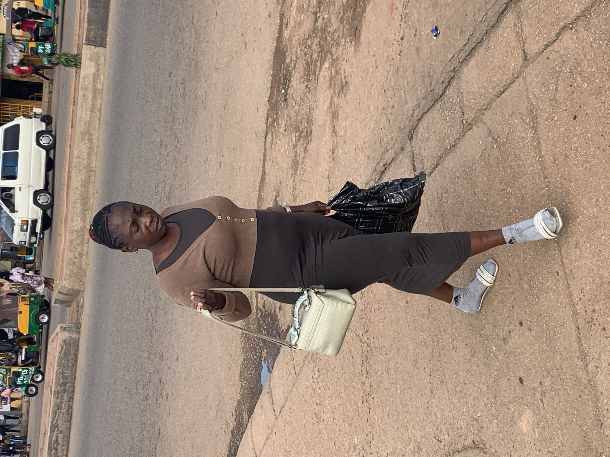 A Black woman, approximately 40-50 years old, walks along a dirt sidewalk in Gura Top, Nigeria. She is wearing a brown cardigan over a dark dress, gray socks, and white sandals. She carries a light-colored purse on her left shoulder and a black plastic bag in her right hand. In the background, a white van and a three-wheeled vehicle are visible on a paved road. The environment appears to be a busy street with various people and vehicles.