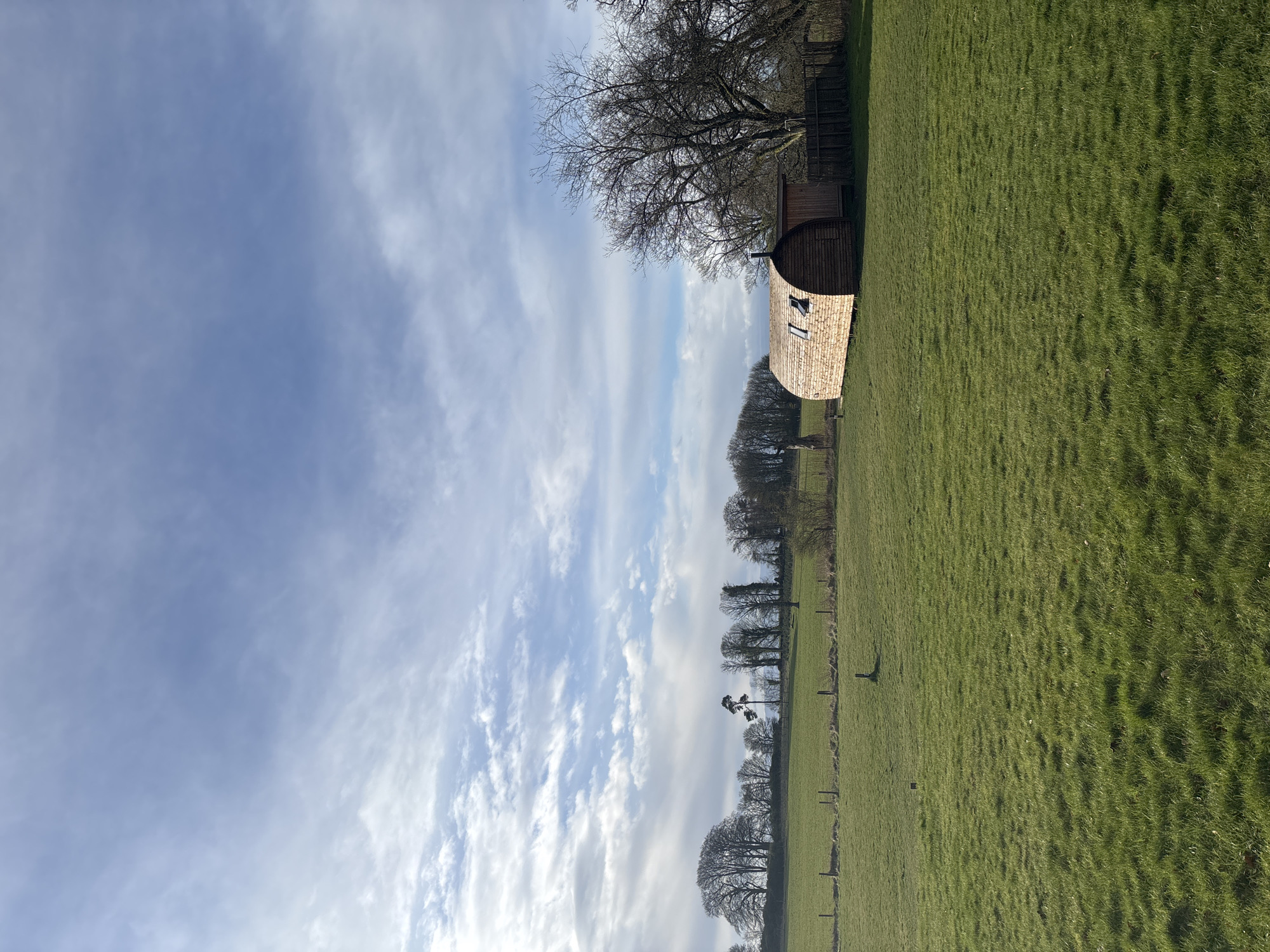 The image presents a view of a rural landscape under a blue, partly cloudy sky. The location appears to be in a field, likely in the Leven, United Kingdom area. The field is covered in green grass. A small wooden structure with a distinctive rounded roof is situated in the upper part of the image, possibly a shed or a small cabin. A tall bare tree stands nearby. In the background, a row of trees delineates a boundary or horizon. Fence posts are visible scattered across the field.