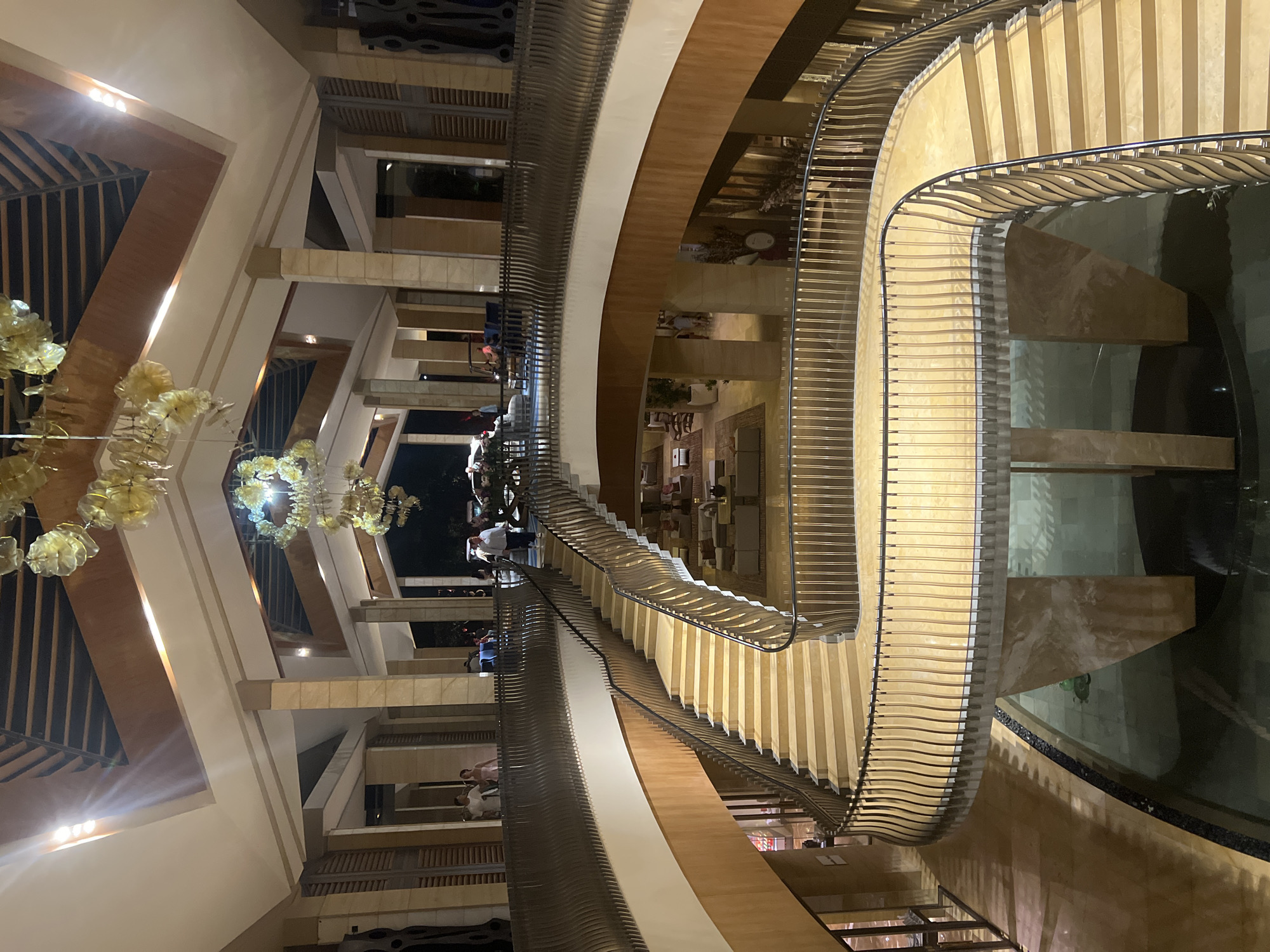 Captured from a high-angle, this photograph shows the interior of a multi-story building, featuring a winding staircase with metal railings. The building’s architecture incorporates elements of wood and stone.
Along the staircase, several individuals can be seen standing or walking.
A notable feature of the scene is a large, decorative chandelier hanging from the ceiling.
The image was taken in Kuta Selatan, Indonesia.