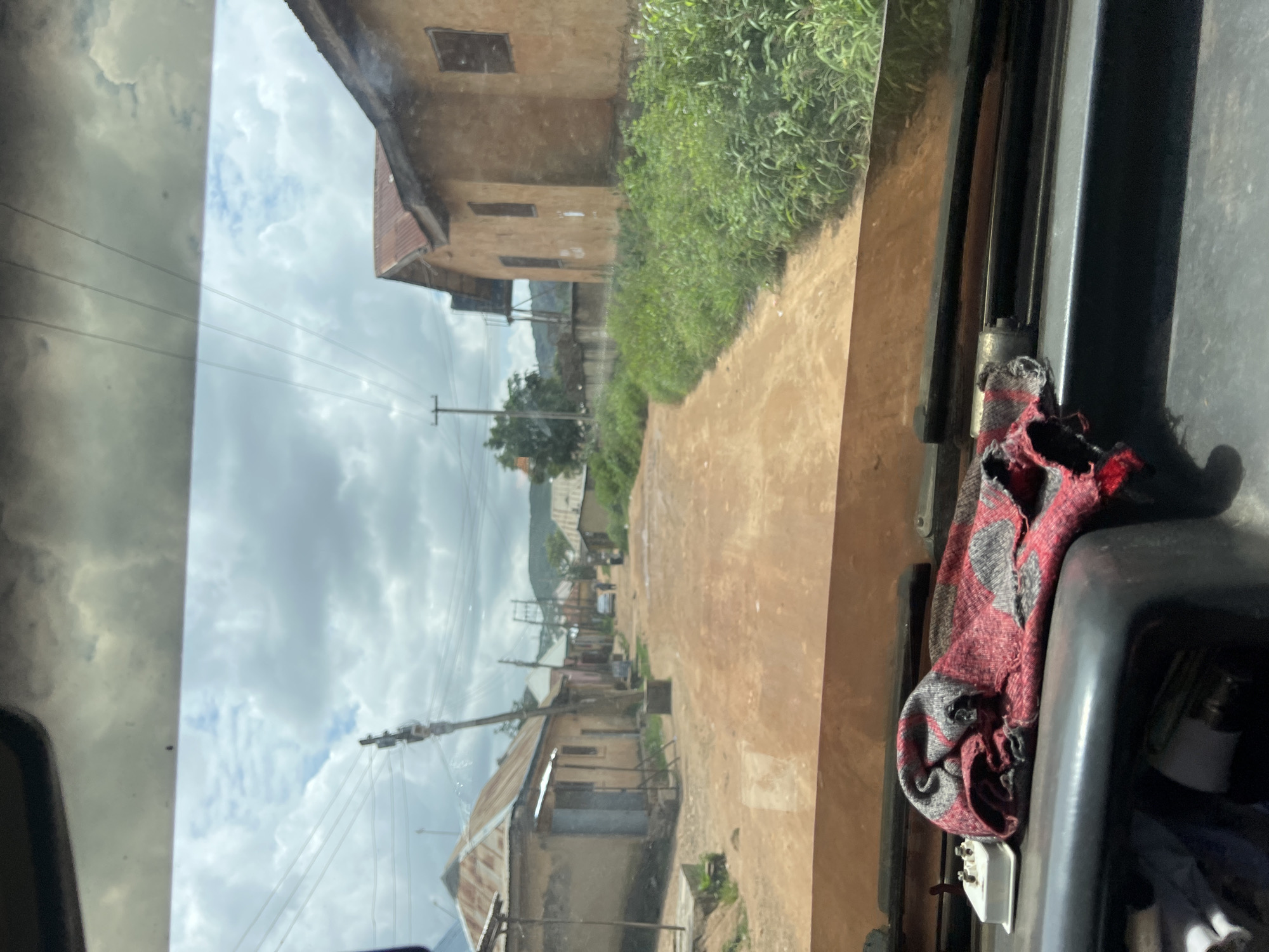 The image, taken from inside a vehicle looking through the windshield, depicts a scene along a dirt road in the city of Rukuba Road, Nigeria. The road is unpaved and appears to be the main thoroughfare of a settlement. On either side of the road, there are simple, weathered buildings constructed with what appears to be mud or unpainted plaster, some with corrugated metal roofs. Dense green vegetation, including tall grasses and bushes, flanks the roadside, particularly on the right. In the distance, hills are visible under a cloudy sky. Power lines with poles run along the side of the road, indicating the presence of electricity. A few people are seen walking on the road in the mid-ground, and a car is parked further down. In the foreground, resting on the dashboard of the vehicle, is a folded red and black checkered cloth and a white electrical outlet. The overall impression is one of a rural or semi-urban environment in Nigeria, characterized by its unpaved roads and modest structures.
