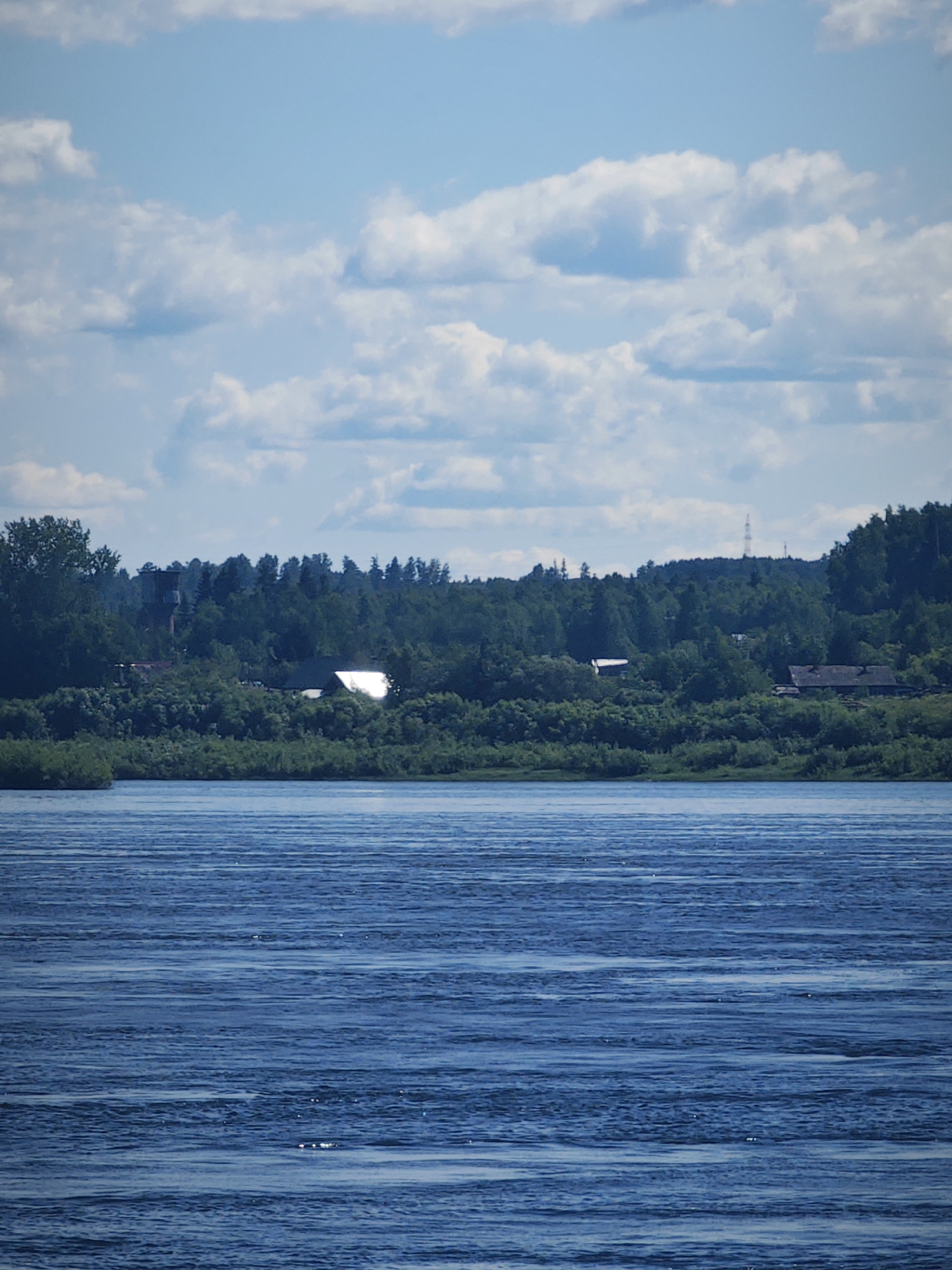 This is a vertical, eye-level, medium shot photograph of a wide, blue river under a partly cloudy sky. The foreground is dominated by the rippling water, which reflects the sky with horizontal lines of light and shadow. In the middle ground, a lush green bank is lined with trees and bushes. Scattered amongst the foliage are several buildings, including houses with pitched roofs and a white structure that gleams in the sun. The background features a dense forest that covers rolling hills, with a faint silhouette of a communication tower visible on the highest peak. The overall impression is one of a serene, rural landscape. The scene is consistent with the village of Абалаково, Russia, situated along the Yenisei River.