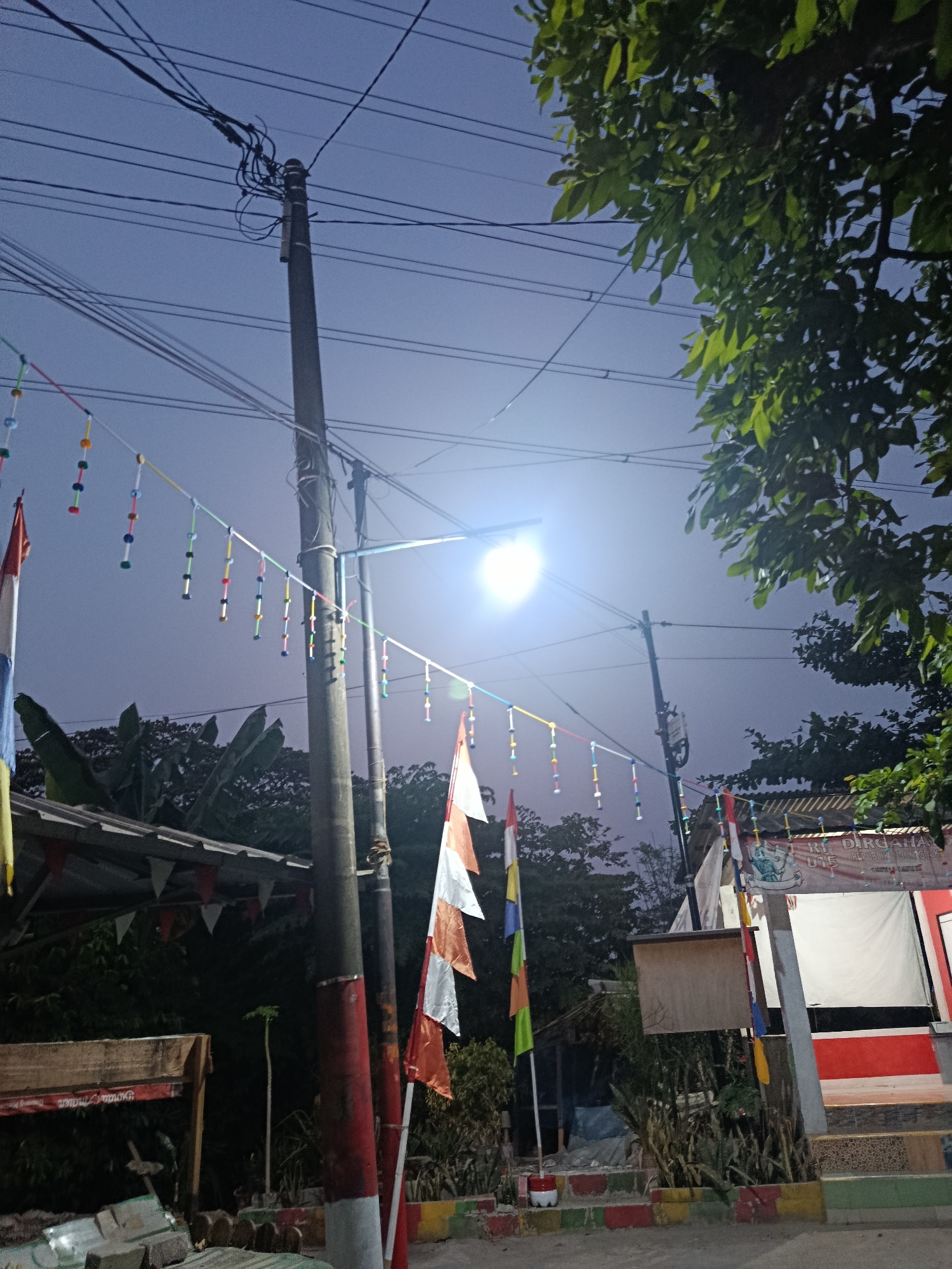 This low-angle, full shot captures an outdoor scene in Wanasari, Indonesia, likely during twilight or early evening, as indicated by the dim ambient light and the illuminated street lamp. The foreground features a section of what appears to be a paved road with a colorful border of red, yellow, and green tiles. To the left, a wooden bench or counter with visible text, possibly in Indonesian, is partially visible. Adjacent to this, a pile of materials, including what seem to be bricks and possibly cement bags, are stacked.
Dominating the center of the image is a tall utility pole, marked with red and white bands near its base, supporting a complex network of electrical wires against the dusky sky. Strung between utility poles and across the street are decorative strings of colorful beads or small cylinders, punctuated by what appear to be Indonesian flags (red and white) and colorful pennant banners hanging from a corrugated metal roof structure on the left.
In the background, the silhouettes of trees and vegetation are visible, with lush green foliage on the right side of the frame, partially obscuring the sky and wires. Several more utility poles are scattered throughout the scene, some with visible wires. On the right, a building with a red and white facade is partially visible, along with a banner that reads "RT DIRGAHA..." and other Indonesian text. The overall impression is a residential or semi-commercial street decorated for a local celebration or event, with elements of everyday infrastructure intertwined with festive adornments. No people are visible in the frame.