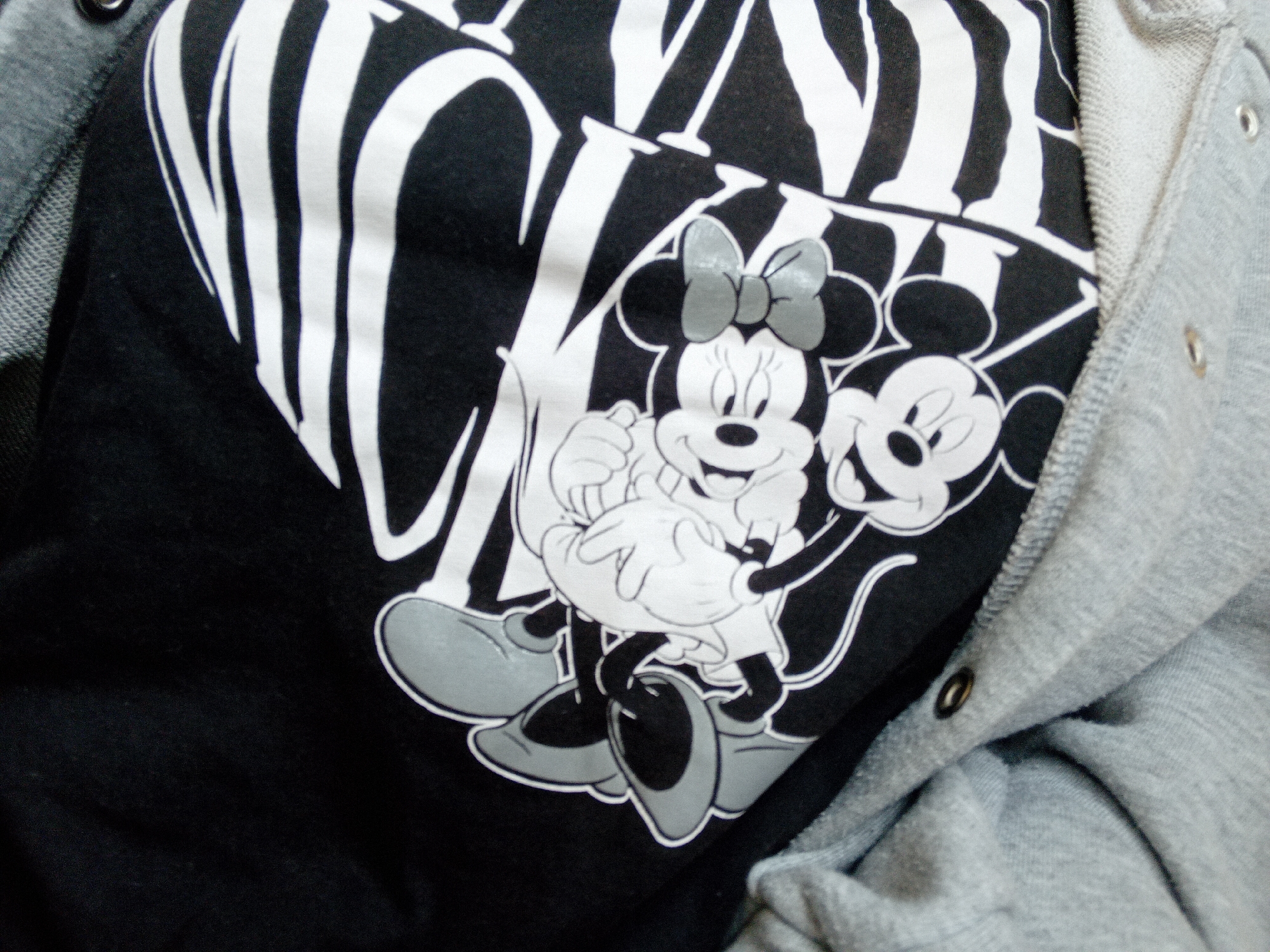 This is a close-up, high-angle shot of a grey jacket or hoodie partially covering a black t-shirt with a graphic print. The print features cartoon characters, appearing to be Mickey and Minnie Mouse, rendered in black and white with grey accents. They are depicted in a playful pose. The grey fabric has visible stitching and snaps. The background is dark and out of focus, suggesting the t-shirt and jacket are the primary subjects. The image was captured in Zuberec, Slovakia.