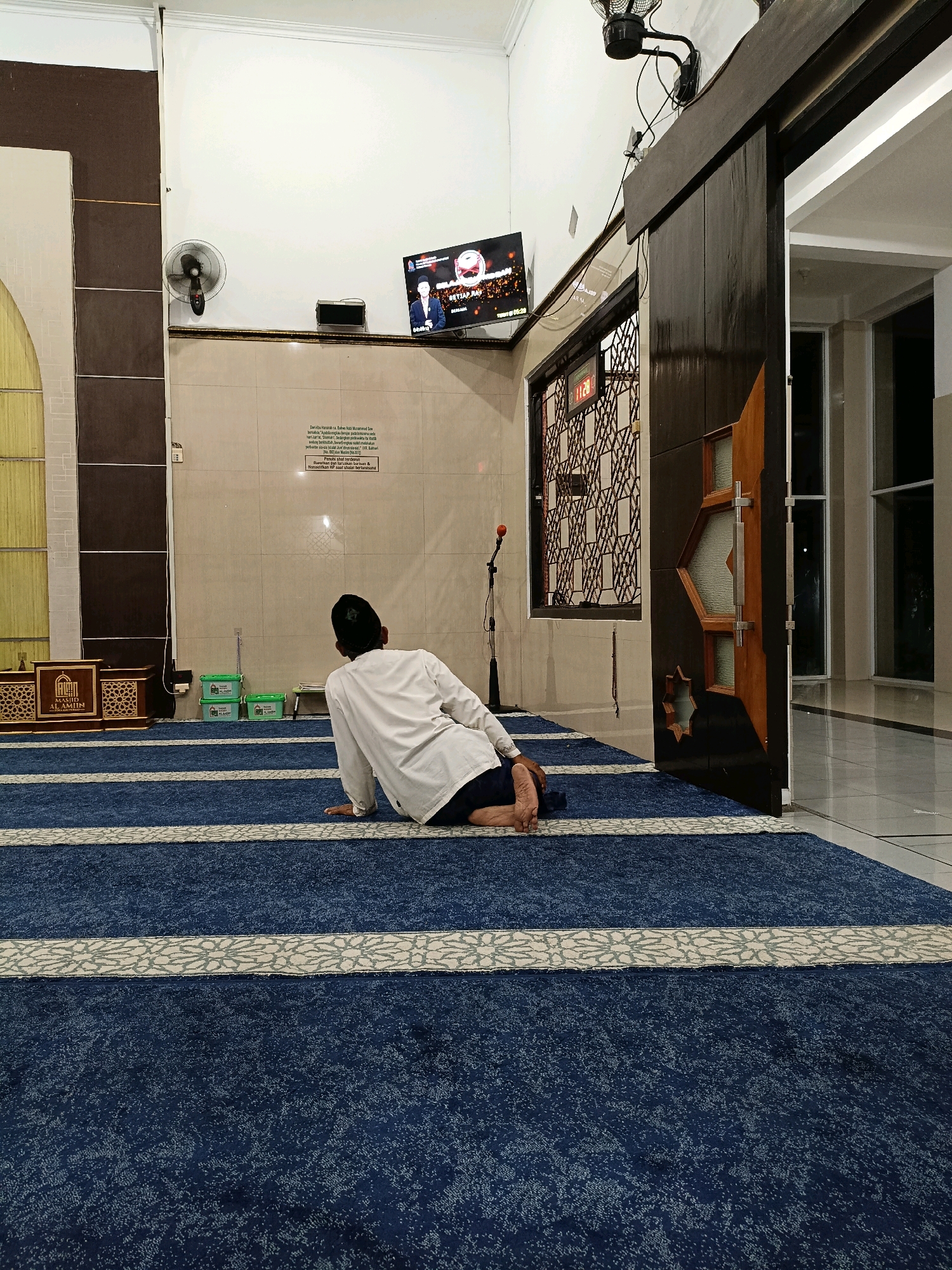 A person wearing a white long-sleeved shirt and a black cap is sitting on a blue carpeted floor in what appears to be a mosque in Maguwoharjo, Indonesia. The person is facing away from the camera, looking towards a television screen mounted on the wall. The television displays a news program with an image of a man in a suit.
The room has beige tiled walls and a dark wooden door with decorative glass panels is open on the right side of the frame, revealing a bright hallway beyond. A fan is mounted on the wall above the television, and a microphone on a stand is positioned in front of the wall. Two green plastic containers are visible on the floor to the left of the person, and a decorative wooden structure, possibly a pulpit or lectern, is also present on the left. The blue carpet has white stripes with a floral pattern. The overall lighting suggests it is indoors, possibly during the evening.