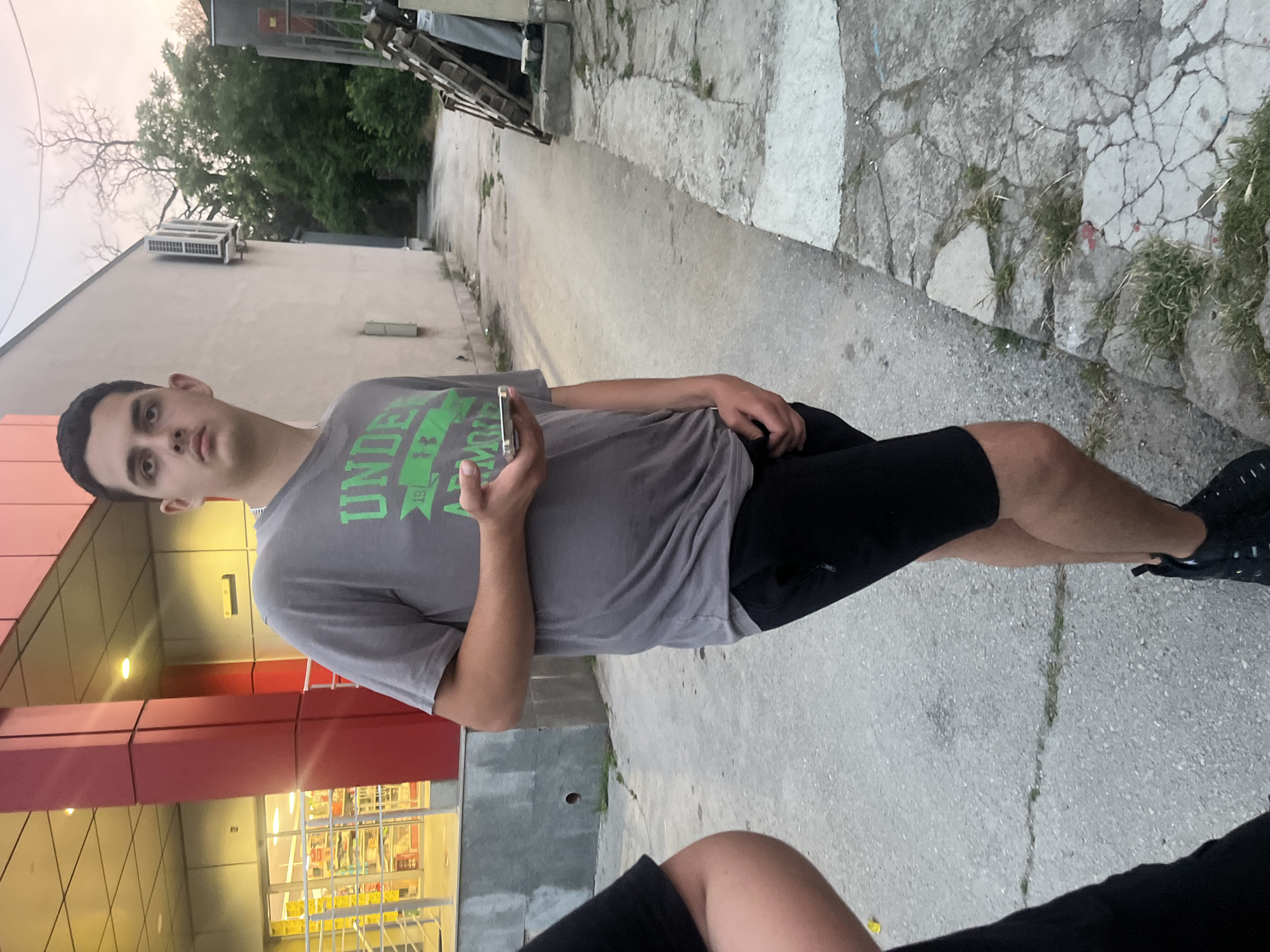 The image captures an outdoor scene in Basarbovo, Bulgaria, depicting a young male standing on concrete pavement in front of commercial buildings, with another individual partially visible on the left.
The central figure is a male, appearing to be in his late teens or early twenties, with short dark hair and fair skin. He is wearing a grey t-shirt with green text, which is partially legible as "UNDER ARMOUR," black athletic shorts, and black athletic shoes. He stands with his body angled slightly towards the right, his head turned to the upper right (viewer's perspective), exhibiting a neutral expression. His left hand holds a silver-colored smartphone, while his right hand is positioned inside the front of his shorts.
To the left of the central figure, the arm and torso of another individual are partially visible, clad in black clothing.
Behind the central figure on the left, a modern-looking commercial building is visible. It features a light-colored facade with prominent red vertical structural elements. A large glass entrance is illuminated from within, revealing shelves stocked with products and yellow safety railings leading up to the entrance.
To the right and further into the background, another building with a plain, light-colored (beige or cream) facade is present. An air conditioning unit is mounted on its wall. Beyond this building, a stack of wooden pallets or similar materials leans against a wall, accompanied by a light-colored fabric or sheet.
The ground consists of grey concrete or asphalt pavement, which is cracked in several places, with small patches of green grass and weeds growing through the fissures. The path extends into the background, appearing to be a narrow alley or side road that curves slightly to the left. In the distant background, beyond the buildings and structures, there are trees with green foliage and some bare, dry branches against a hazy, light-colored sky with subtle pinkish-orange hues.