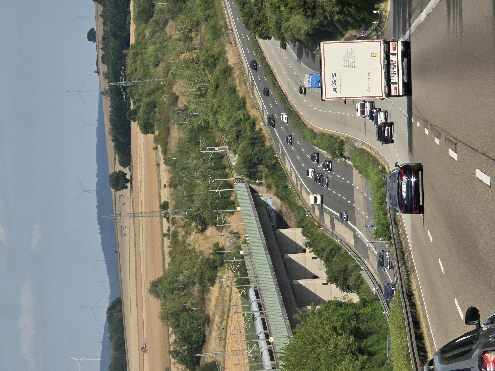 The image depicts a wide, multi-lane highway with several vehicles traveling in both directions. In the foreground, a dark-colored car is visible from the rear, traveling in the right lane. Further ahead, a white semi-trailer truck with a blue trailer is in the left lane, and a white camper van is in the right lane. On the highway to the left, multiple cars are traveling, including a black SUV, a silver sedan, and a white hatchback.
An elevated train track with a modern silver train is visible on a concrete bridge spanning across the highway and the surrounding landscape. The landscape consists of rolling hills covered in green trees and fields of harvested crops, with a few wind turbines visible in the distance on a hilltop. Power lines and electrical towers are also present throughout the scene. The sky is partly cloudy. The image is taken from an elevated perspective, looking down onto the highway and surrounding area. The context of Brechen, Germany, is provided by the presence of a sign with "ASS TRANSPORTE" and a German license plate on the semi-trailer truck, as well as the typical German highway and landscape features.