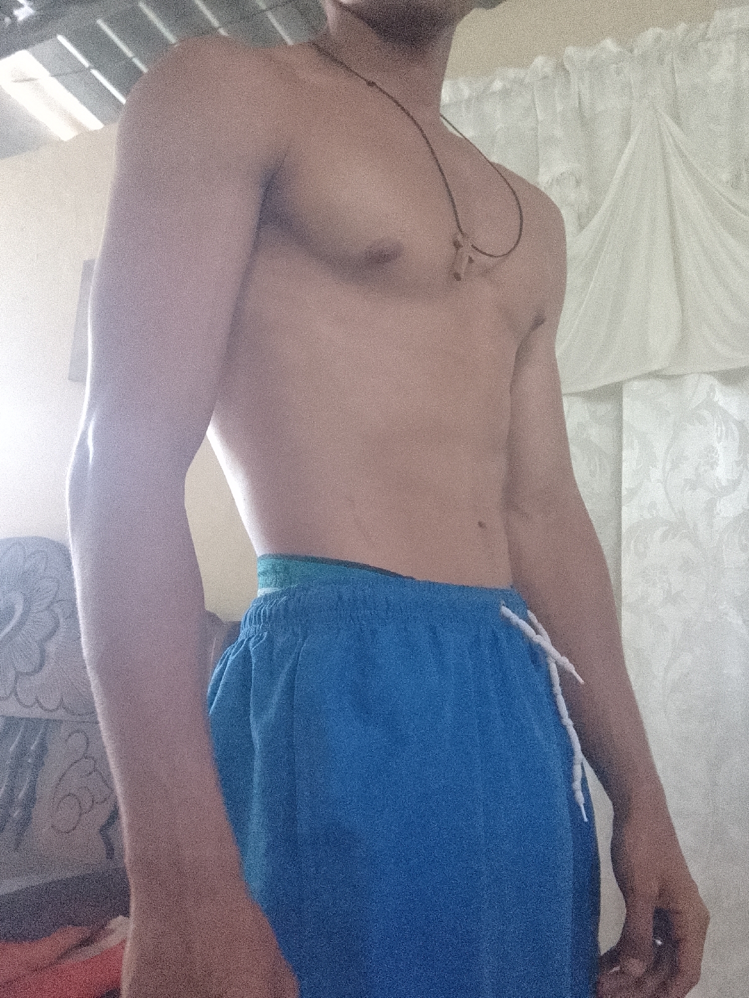 A shirtless individual wearing blue shorts stands indoors. They have a slender physique. A cross necklace is visible around their neck. The background includes a white curtain on the right and what appears to be a decorated chair on the left. The location is suggested to be Juan Griego, Venezuela. Details about the specific building or immediate surroundings are not provided.