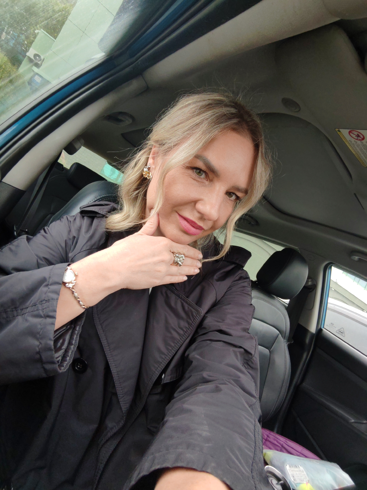 A woman with blonde hair and fair skin is sitting in the driver's seat of a car, taking a selfie. She is wearing a black jacket, earrings, a ring, and a watch. Her left hand is resting on her chin. The car interior is visible, including the seats and the ceiling. The background outside the car is blurred. The lighting suggests it is daytime.