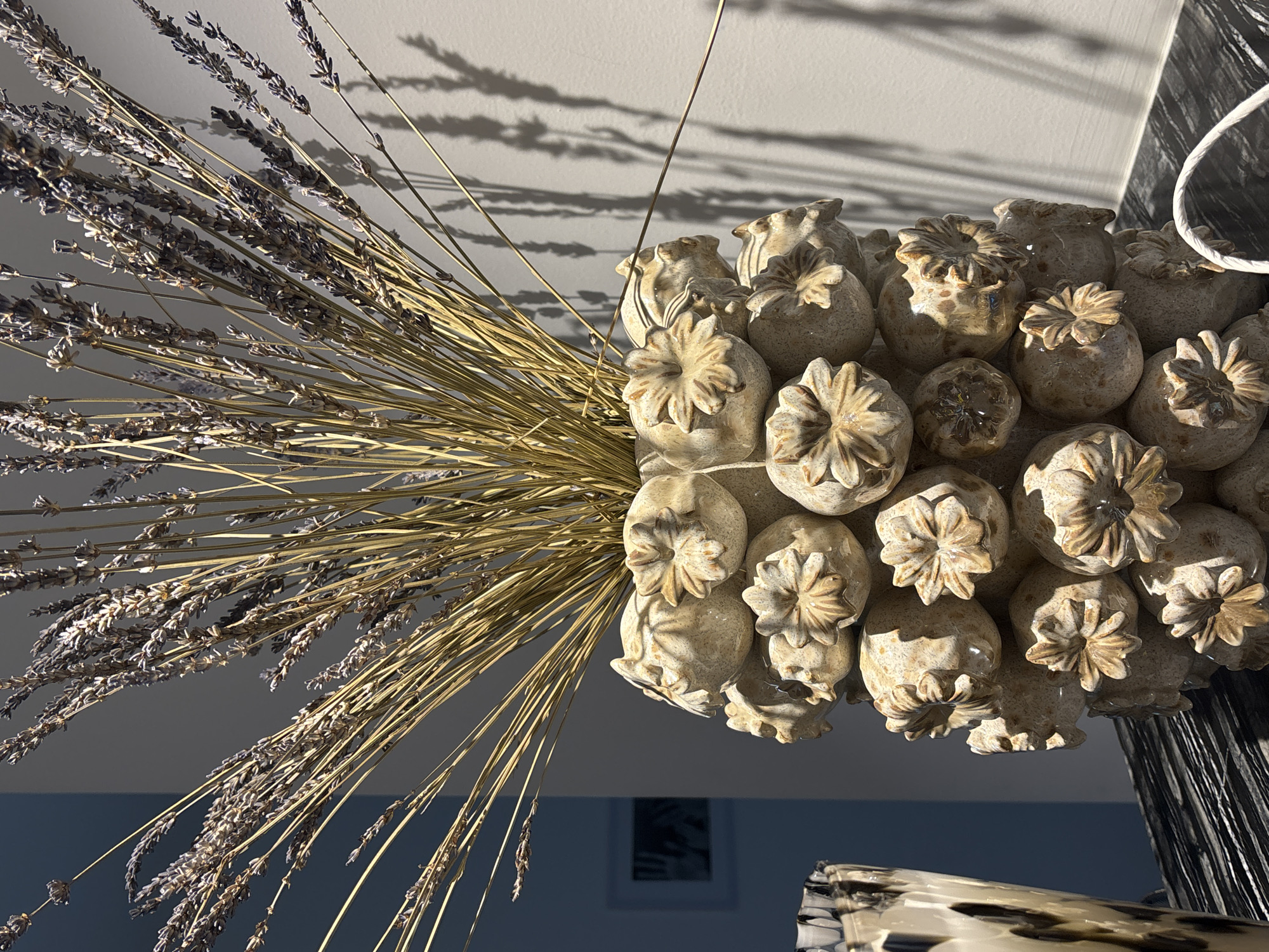 This is a medium close-up, eye-level, indoor photograph of a decorative vase filled with dried lavender. The vase is light beige in color and has a textured surface, appearing to be made of ceramic or a similar material. It is adorned with multiple sculpted poppy seed heads, each with a star-shaped opening at the top. The dried lavender stalks are a muted golden-brown and are arranged in a fanned-out manner, extending upwards and out of the vase. The background consists of a plain, light gray wall, where shadows from the lavender stalks are cast. To the left of the vase, a portion of another vase or decorative object with a black and white marbled pattern is visible. The scene is lit by a light source casting strong shadows, suggesting artificial lighting or direct sunlight from a window. The overall impression is one of still life, focusing on texture and form. There is no indication of the city of Dubai or the United Arab Emirates in the image.