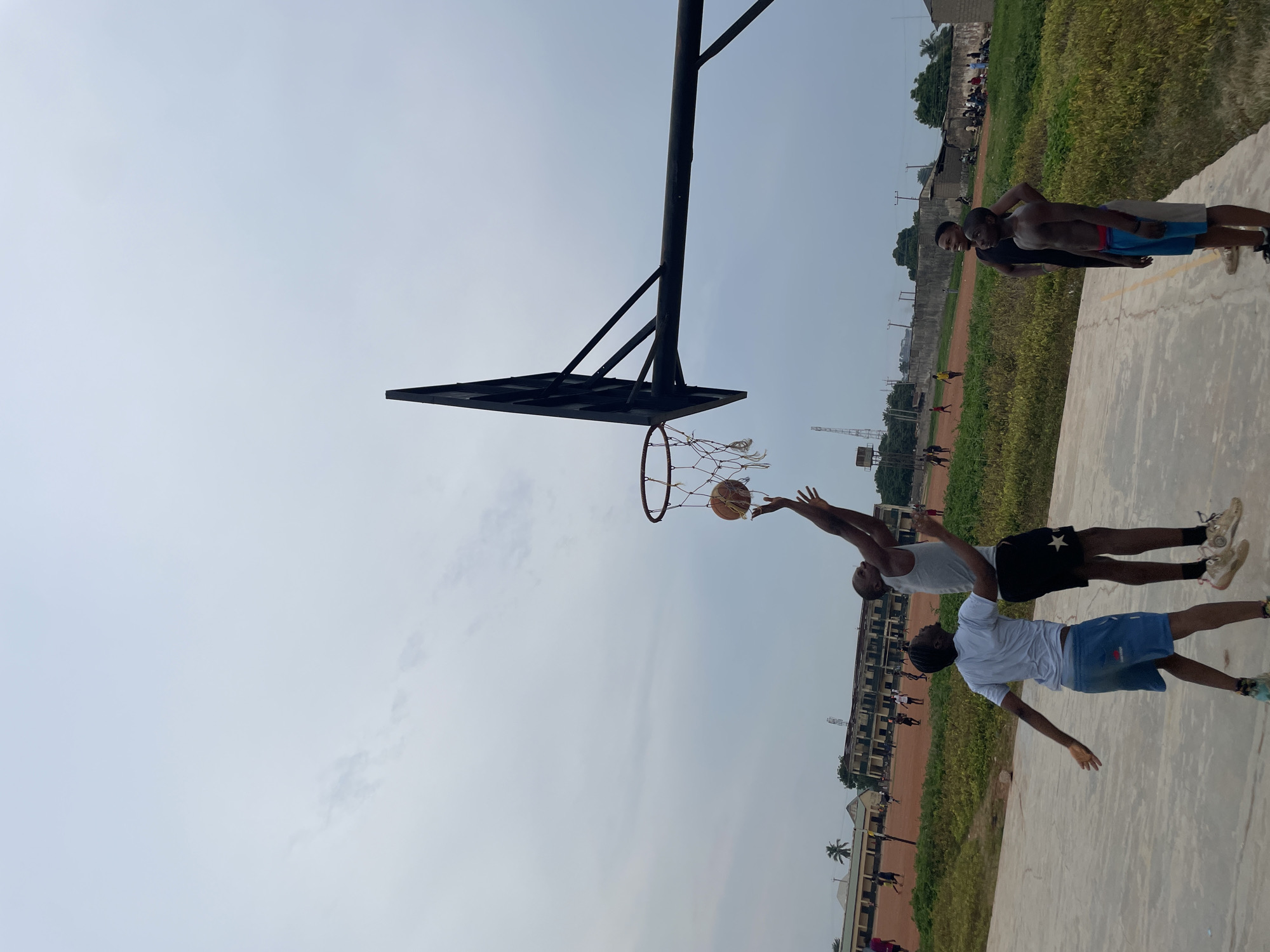 In Akwanga, Nigeria, under a cloudy sky, a basketball game is in progress on an outdoor court. Four young men are actively engaged in the game. One player is mid-air, attempting a shot, while another player is positioned defensively below him. Two other players observe the action, with one standing to the side and the other looking up. The court is made of concrete, bordered by a reddish-brown dirt area. In the background, there are buildings, including what appear to be school structures, and lush green vegetation. The overall scene depicts a casual and energetic basketball match in a local setting.