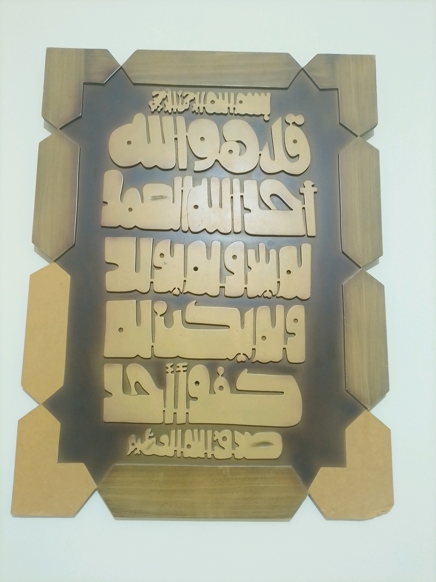 The image shows an Islamic calligraphy artwork displayed on a white wall. The artwork is framed with wood and features Arabic calligraphy in a stylized font. The calligraphy appears to be made of a lighter colored material, possibly wood or metal, and is raised from a dark background. The overall impression is of a decorative and religious piece. The image is a medium shot, focused on the artwork itself, with no other objects or people visible. The lighting is even, suggesting it was taken indoors with artificial light. There is no information in the image to suggest a location in Amman, Jordan.