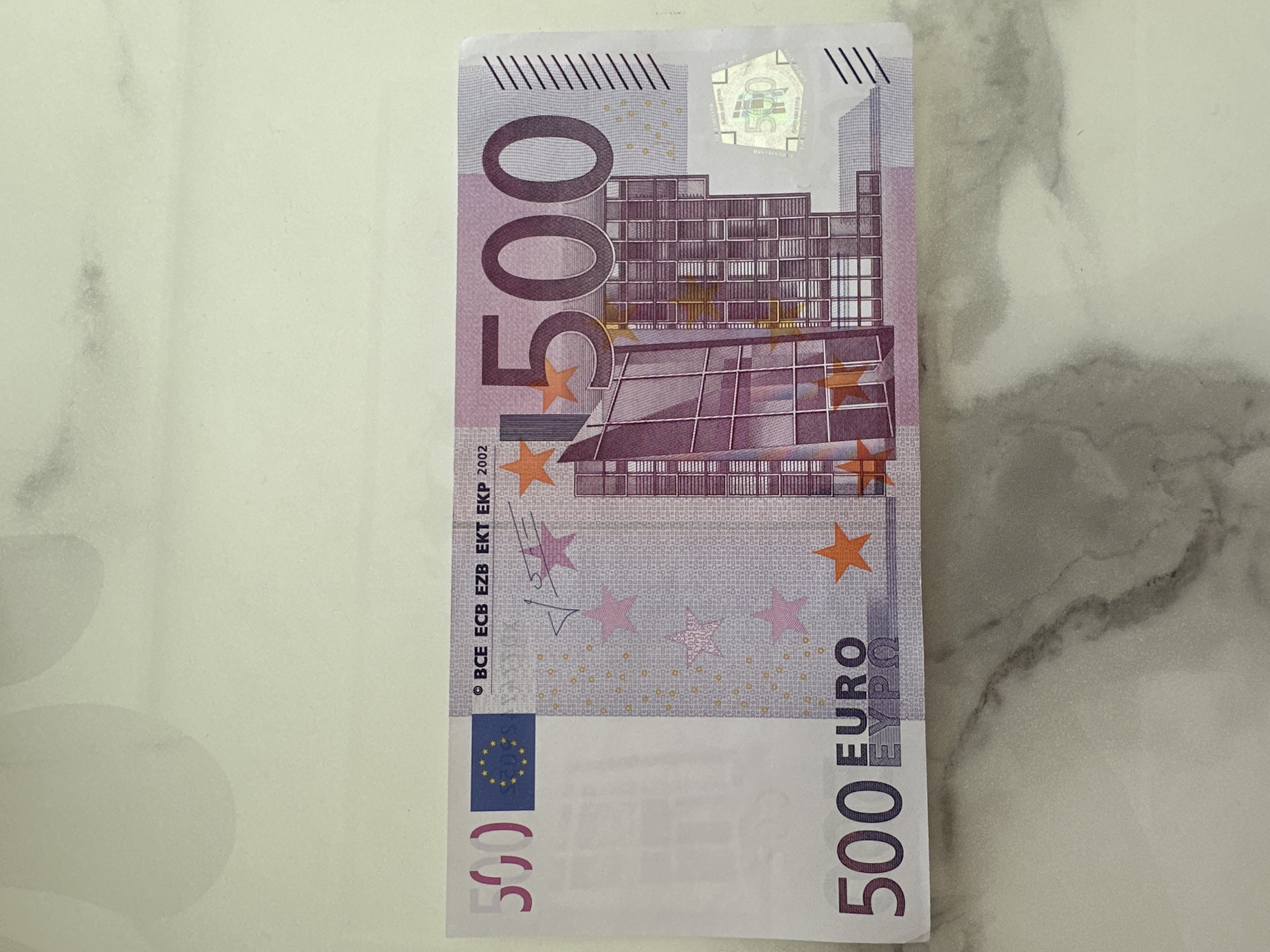 A single 500 Euro banknote, series 2002, is depicted face-up on a light-colored, subtly veined marble or stone surface. The banknote, primarily purple and lilac, displays the European Union flag and a "500" watermark in the upper left corner. Large "500" numerals are visible on the upper right and lower left, alongside "500 EURO EYPΩ" at the bottom left. The design features a modern architectural motif dominating the right half, complemented by orange stars. A holographic patch showing a "500" and architectural elements is situated on the right side. Microprinting is integrated throughout the note's design. The banknote appears flat and well-preserved. The background surface is smooth and reflects diffuse, overhead lighting.