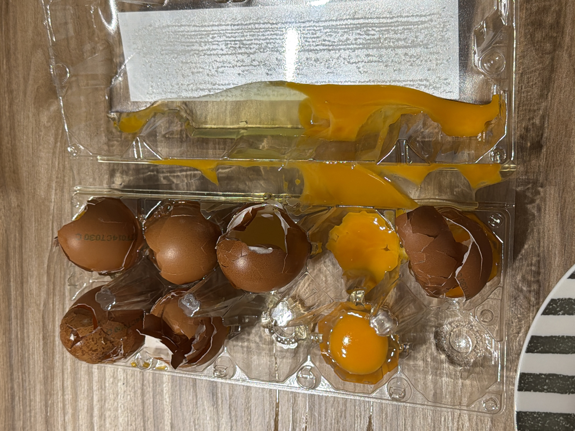 A carton of eggs, several broken with yolk and white spilled within the plastic container. The carton is on a wooden surface. The location is unspecified and Catania, Italy is not evident from the provided image.