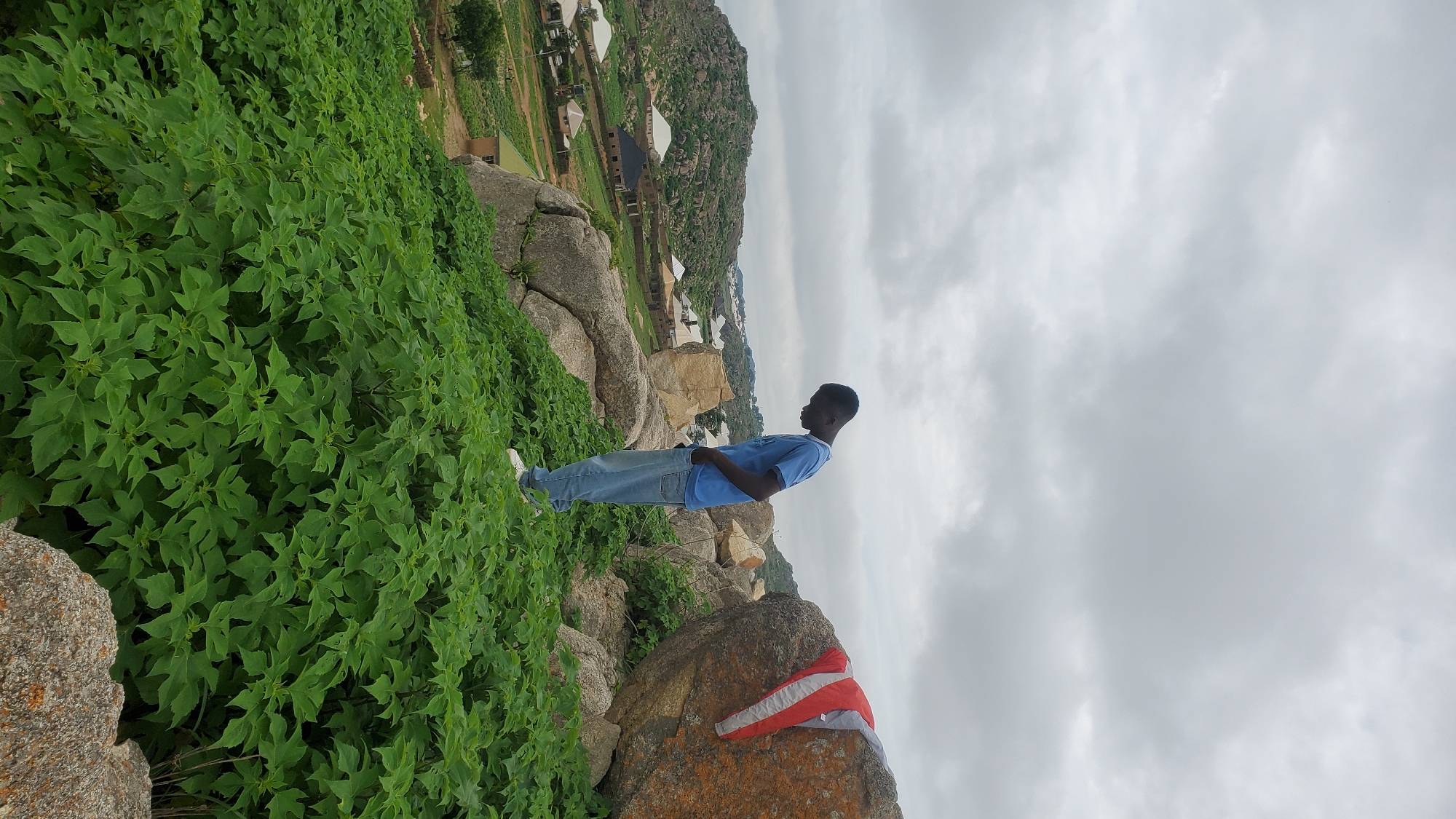 A young Black man wearing a blue t-shirt and blue jeans stands on a rocky, overgrown hillside overlooking a settlement in Jos, Nigeria. He is looking down, with his hands in his pockets. Behind him, a large boulder has a red, white, and gray jacket draped over it. The settlement below consists of various houses with different colored roofs, surrounded by greenery and rocky outcrops. The sky is overcast with gray clouds.