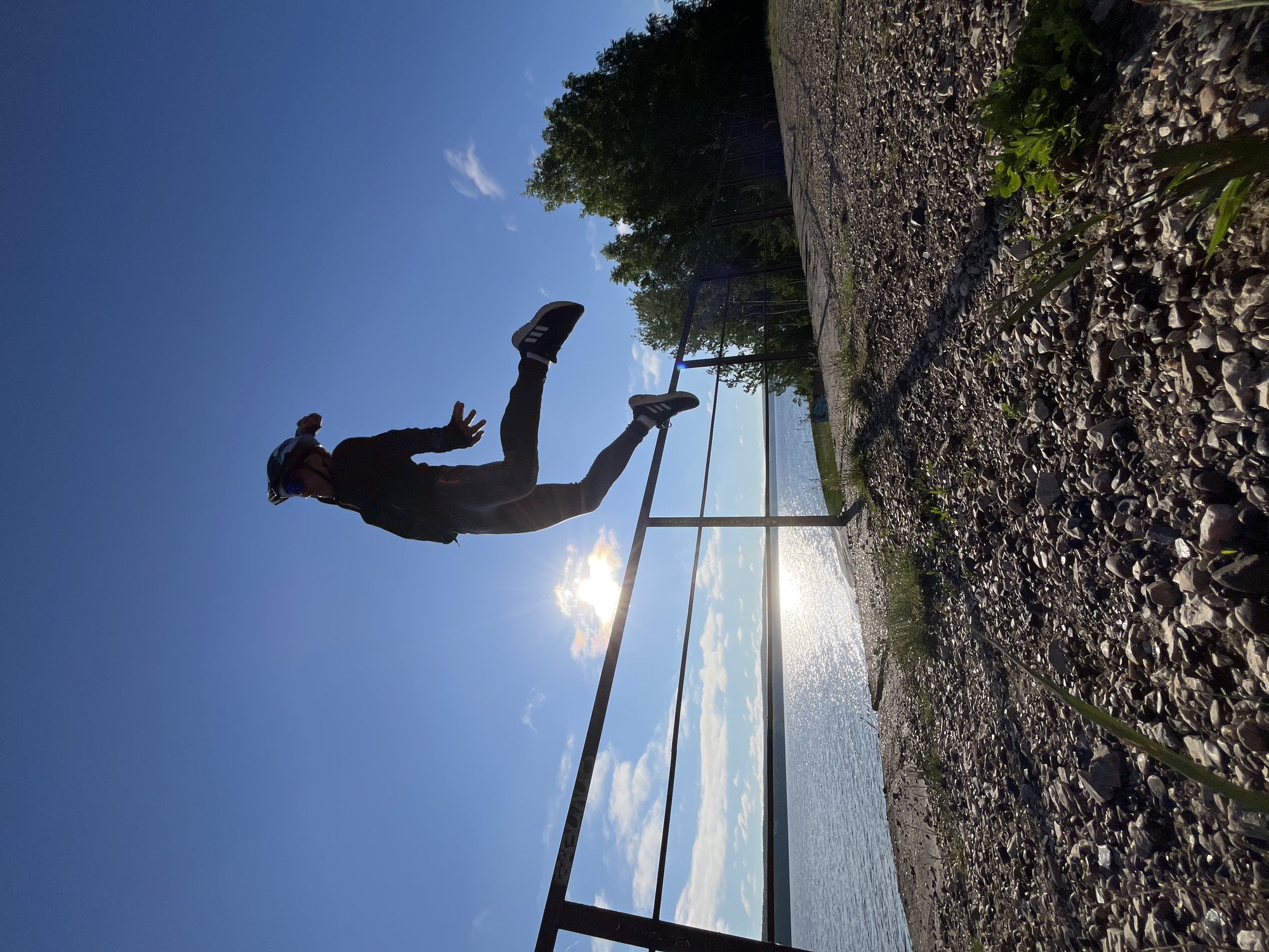 A person in silhouette is captured mid-jump over a railing next to a body of water. The individual is wearing a helmet and dark athletic clothing, with one leg extended forward and the other bent. The sun shines brightly behind the person, casting a glare on the water and creating lens flare. The railing is a dark, metallic structure running horizontally across the frame. The foreground consists of a path covered in pebbles and grass. In the background, lush green trees line the edge of the water. The sky is a clear, vibrant blue with a few scattered clouds. This scene likely depicts the embankment area of the Kama River in Chaikovsky, Russia, a popular location for outdoor recreation.