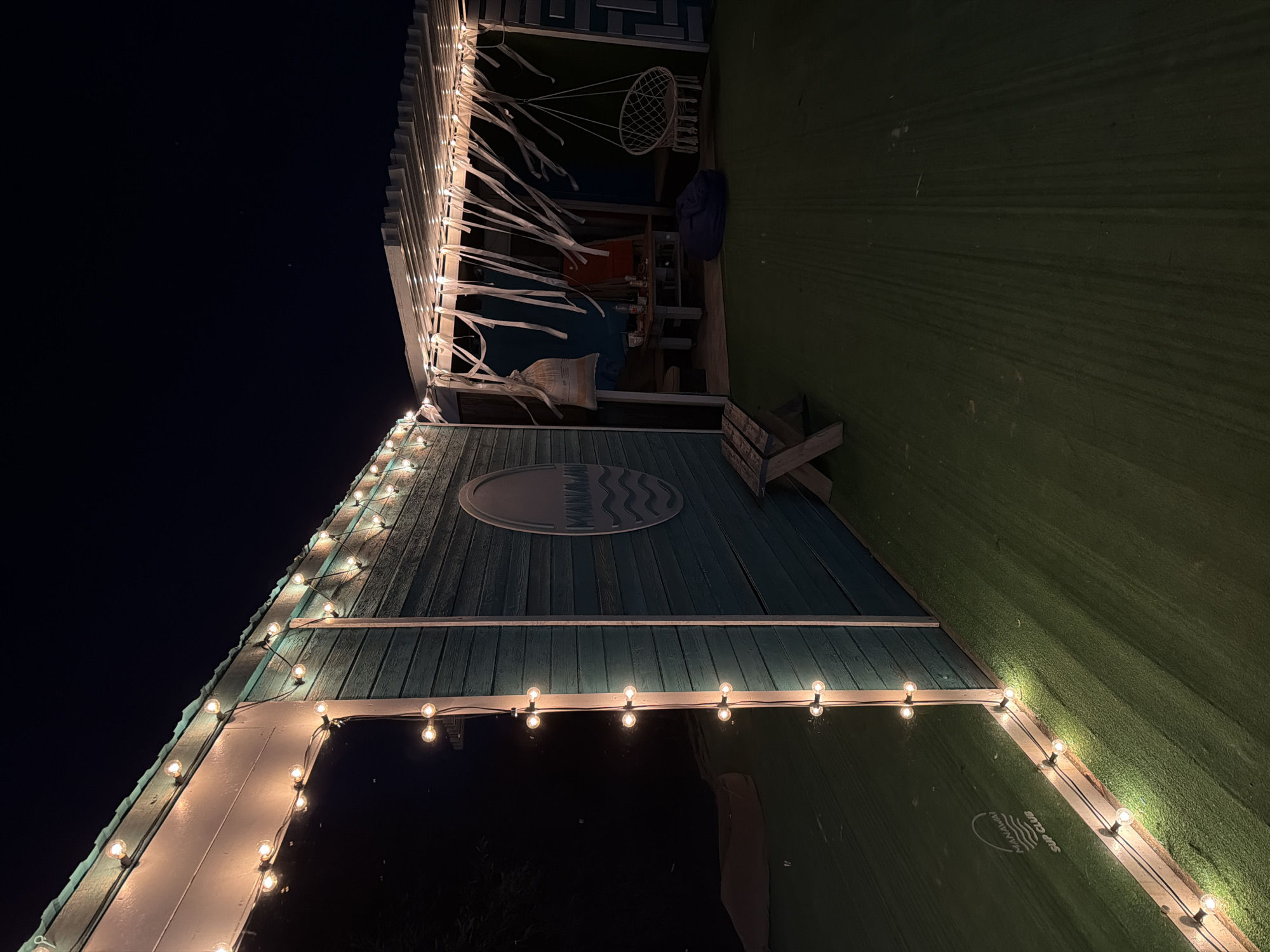 An outdoor nighttime scene depicts a teal-colored wooden structure with a sloped roof adorned with a string of warm-toned lights. The structure has a large, reflective window on the left side, revealing a dark, indistinct interior. A circular emblem with wavy lines and text that reads "MANAWA SUP CLUB" is affixed to the exterior wall.

Adjacent to the structure is a covered patio area with a slatted roof, also illuminated by string lights. White streamers hang from the patio ceiling. Within the patio, a white macrame hanging chair is suspended. A dark blue beanbag chair rests on the green, artificial turf ground. A wooden picnic table with benches is visible further back, with bottles and other objects on its surface. A small, wooden bench sits in front of the teal structure. The overall environment suggests a recreational or gathering space. The location is identified as Otradnoye, Russia.