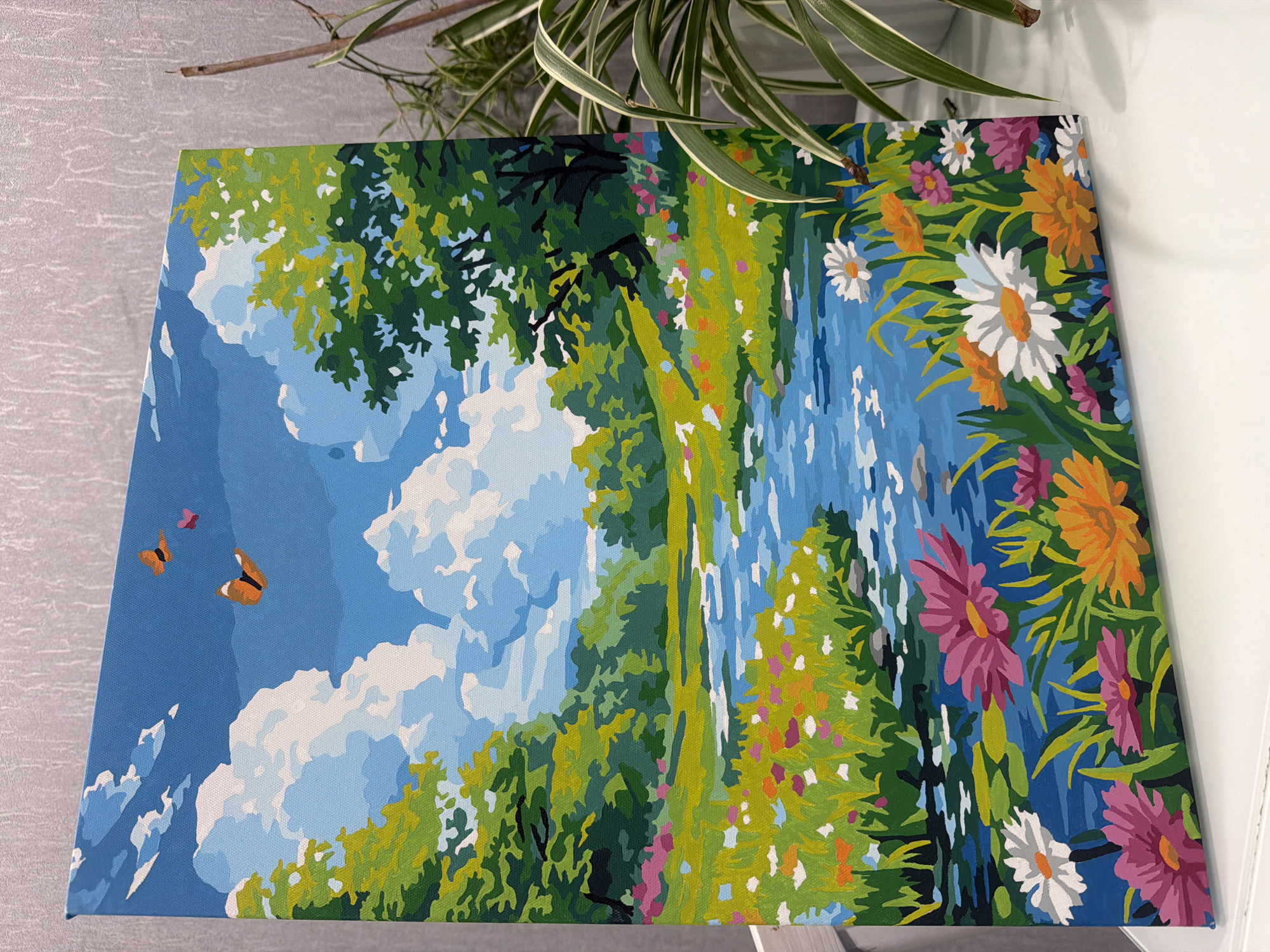 The image displays a vibrant, cartoon-style landscape painting, rendered in a paint-by-number aesthetic on a stretched canvas. The upper portion features a bright blue sky with stylized white clouds, two orange butterflies, and a single pink heart-shaped object in flight. A meandering blue river or stream flows through the center, displaying lighter blue ripples and reflections, with some green lily pads visible. Its banks are adorned with lush green grass and numerous colorful wildflowers, including white daisies, pink and purple asters, and orange blossoms. The background consists of dense green foliage and trees, suggesting a gentle, rolling landscape.
The painting is displayed at an oblique angle, leaning against a textured light gray wall with a subtle pattern. It rests on a flat, white surface. To the right of the painting, a houseplant with variegated green and white leaves is partially visible, its foliage extending into the frame. There are no people visible, and no actions or interactions beyond the depicted butterflies in the painting. There are no discernible details within the image to indicate its specific location in Rostov-on-Don, Russia, or any other geographic area.