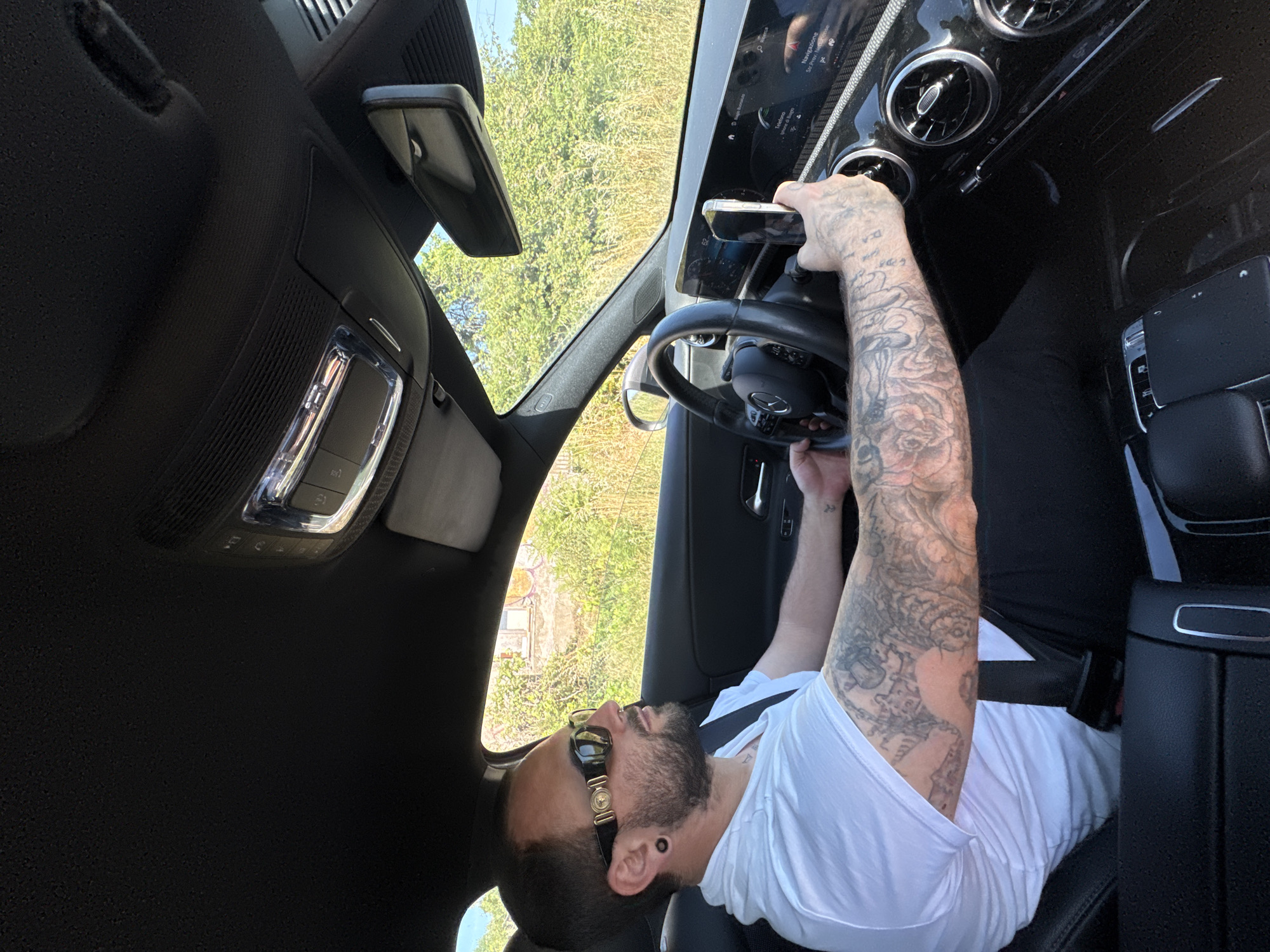 The image shows a man seated in the driver's seat of a car, possibly a Mercedes-Benz based on the steering wheel emblem. He has a beard, is wearing sunglasses, and has extensive tattoos on his left arm. He is holding a smartphone in front of the car's infotainment screen. The environment visible through the windshield suggests an outdoor setting with greenery. It is impossible to independently confirm the location of Nettuno, Italy.