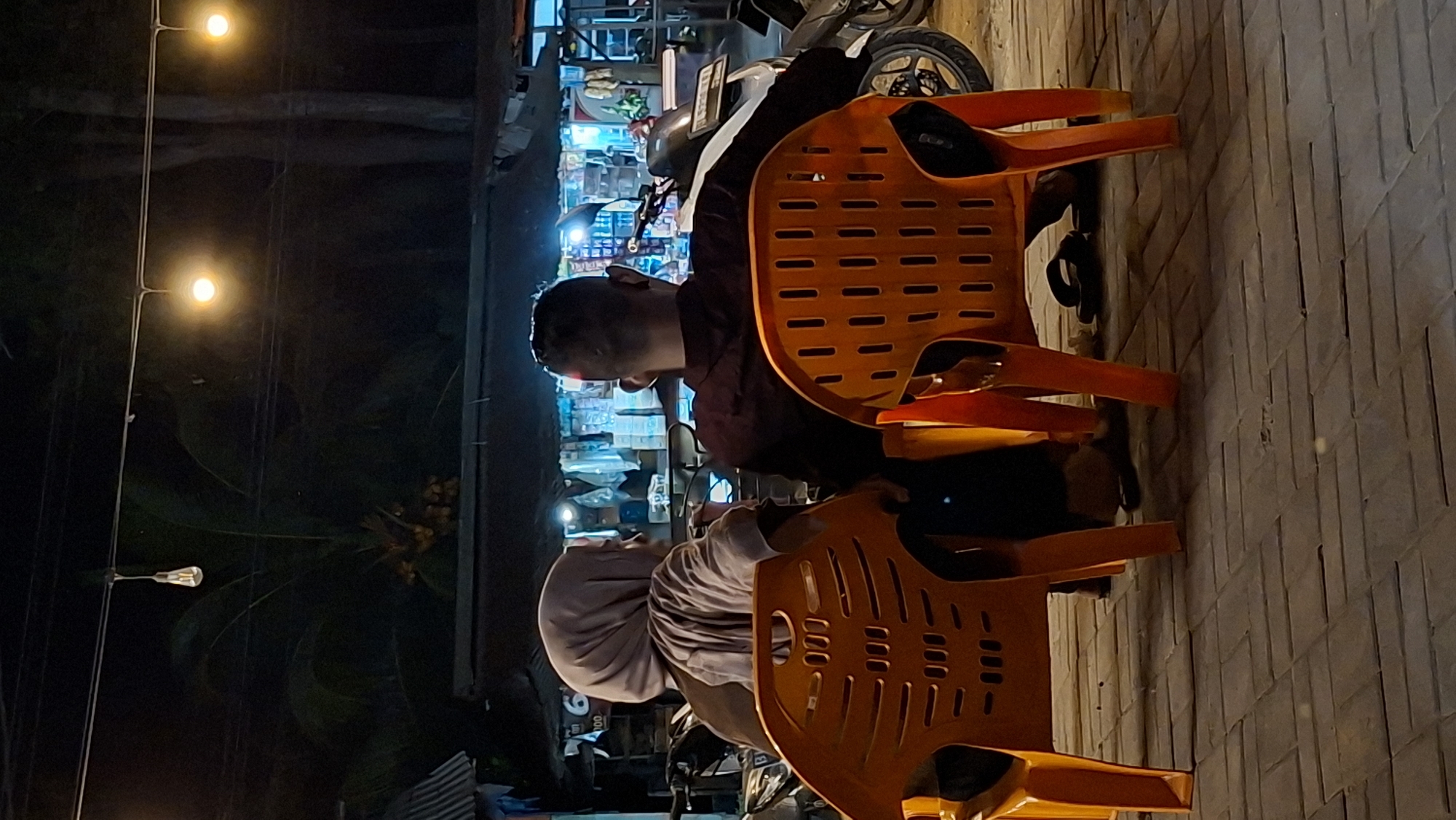 The image depicts two individuals, a man and a woman, seated on orange plastic chairs in an outdoor setting at night. The man is wearing a dark shirt and is looking away from the camera, his profile visible. The woman is wearing a light-colored hijab and is also looking away. They appear to be at a roadside stall or small shop, indicated by shelves with various items behind them. A motorcycle is parked nearby. The ground is paved with rectangular tiles. Two warm-toned lights hang from wires above, illuminating the scene. The overall atmosphere is casual and seems to capture a moment of rest or conversation. The time of day is clearly night. The location is identified as Keudeteupinpunti, Indonesia. There is no visible text in the image.