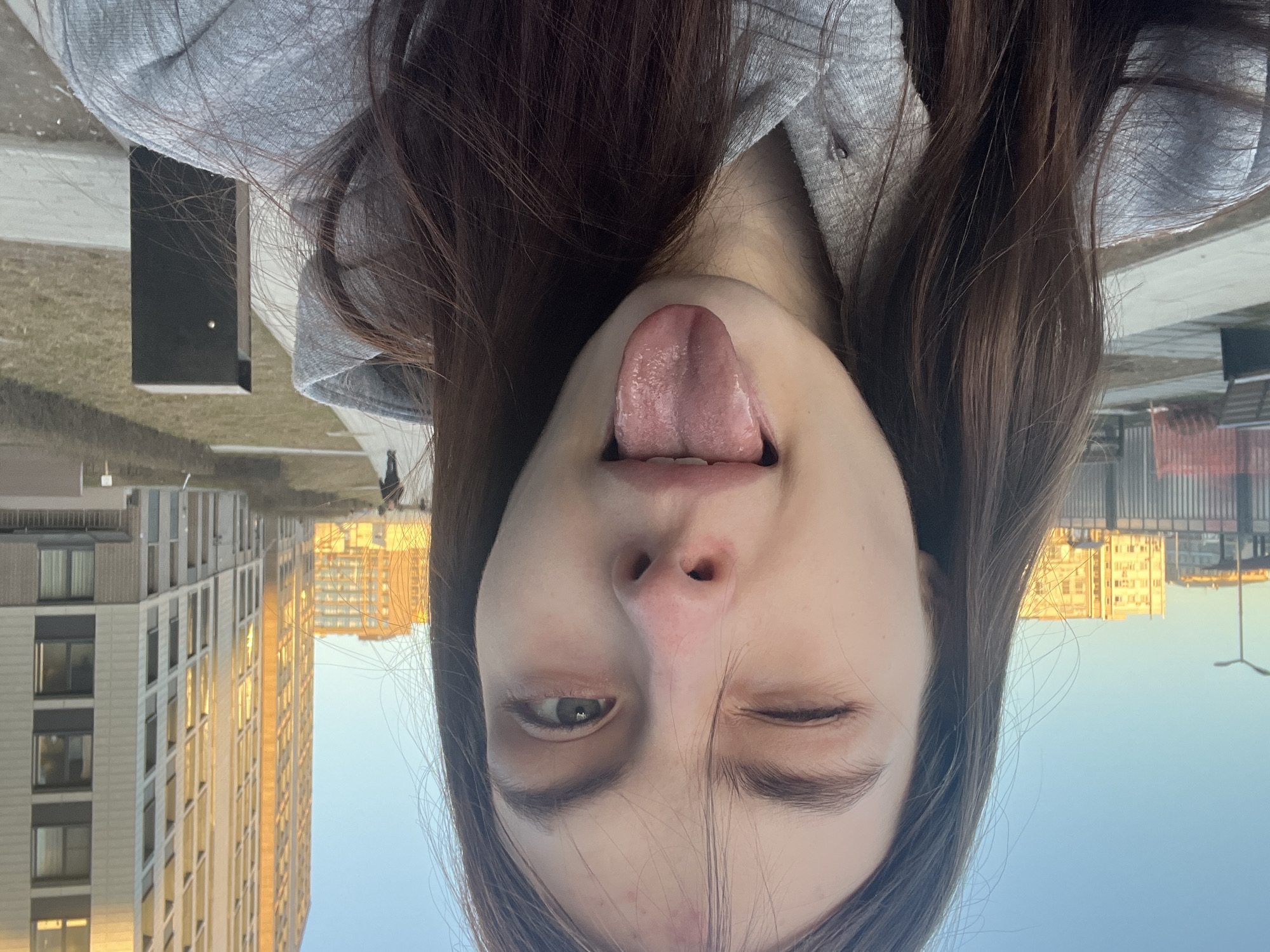 The image is a selfie taken by a young woman with long, dark hair, wearing a gray hooded sweatshirt. She is upside down in the frame and sticking her tongue out while winking one eye.
The background reveals an urban environment. On the left side of the frame, there is a light-colored high-rise building with multiple windows. In the background, a building reflects the setting sun. There is a small black rectangular structure and a figure of a person in the distance. The sky is a clear, pale blue.
The location is likely in Saint Petersburg, Russia, based on the architecture of the buildings.