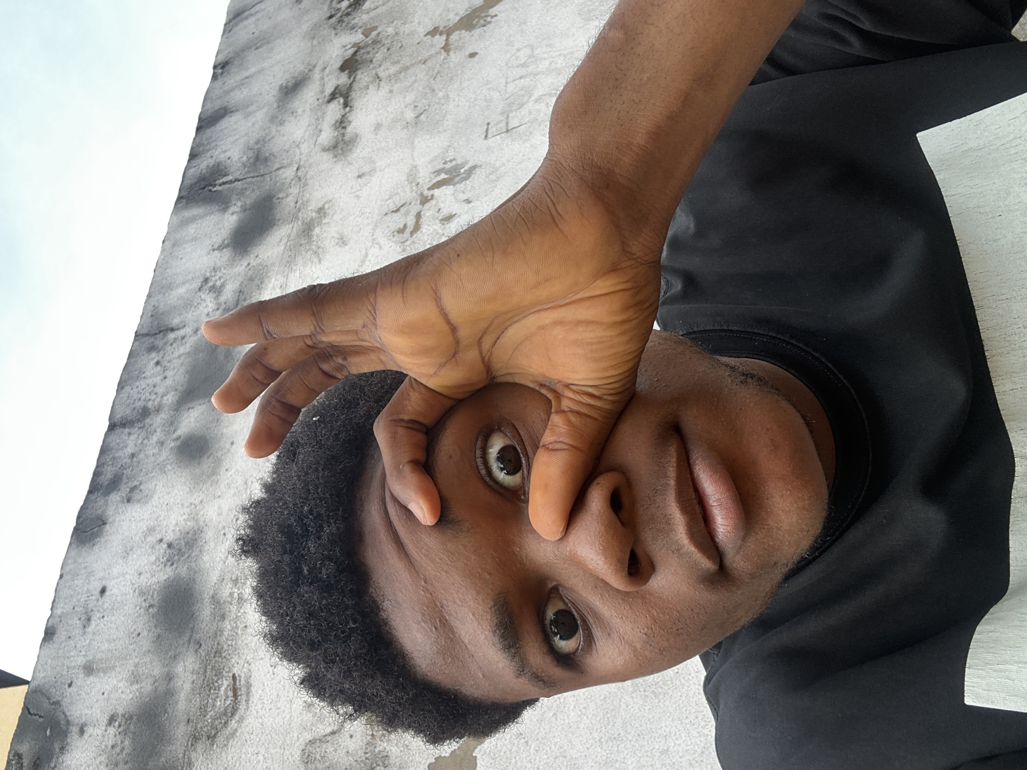 A close-up, eye-level, outdoor medium shot features a young Black person wearing a black t-shirt with a white rectangular design across the chest. The person is looking directly at the camera, with their right hand raised and the thumb and index finger forming a circle around their right eye. Their left eye is partially obscured by their hand. The person's hair is short and dark, with a curly texture. Behind the person is a rough, textured concrete wall that appears weathered and stained, with patches of shadow. The sky is visible in the upper background. The scene appears to be set in Ihiagwa, Nigeria, given the architecture and overall environment.