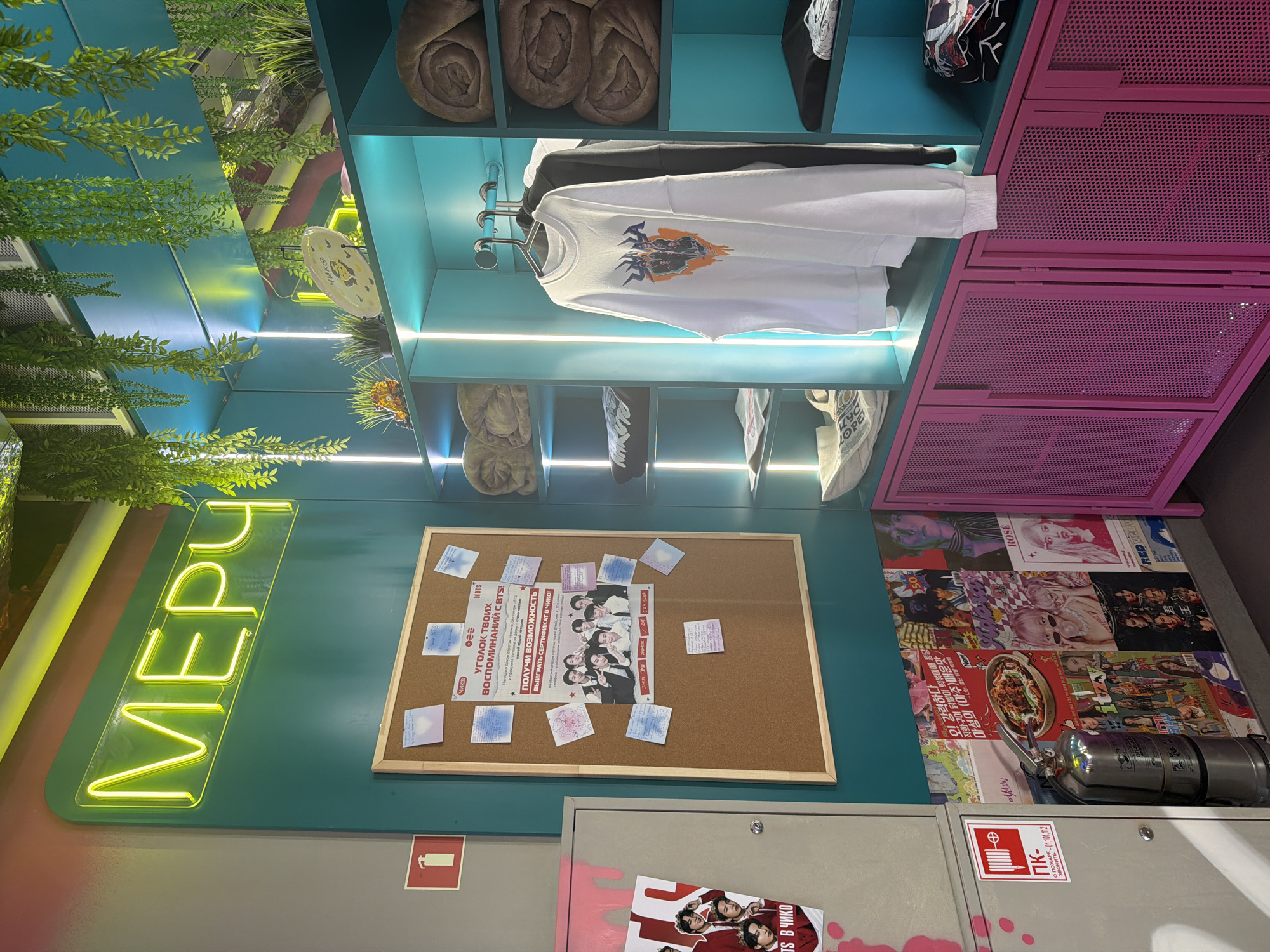 The image shows a corner of a room, possibly a store, in Perm, Russia. On the top left, there are artificial green plants hanging from the ceiling. Below, a shelf unit is visible, with items like rolled-up blankets, clothing, and a white sweatshirt with a printed design. To the right of the shelves, there is a pink, perforated metal cabinet.
On the wall, there is a neon sign that reads "МЕРИ". Below the sign, there is a corkboard covered with paper notes and images, a fire extinguisher and a poster. The corkboard also has a newspaper with a headline mentioning "BTS". Underneath is a metal cabinet box. The floor has a collage of images that appear to be magazine clippings or posters.