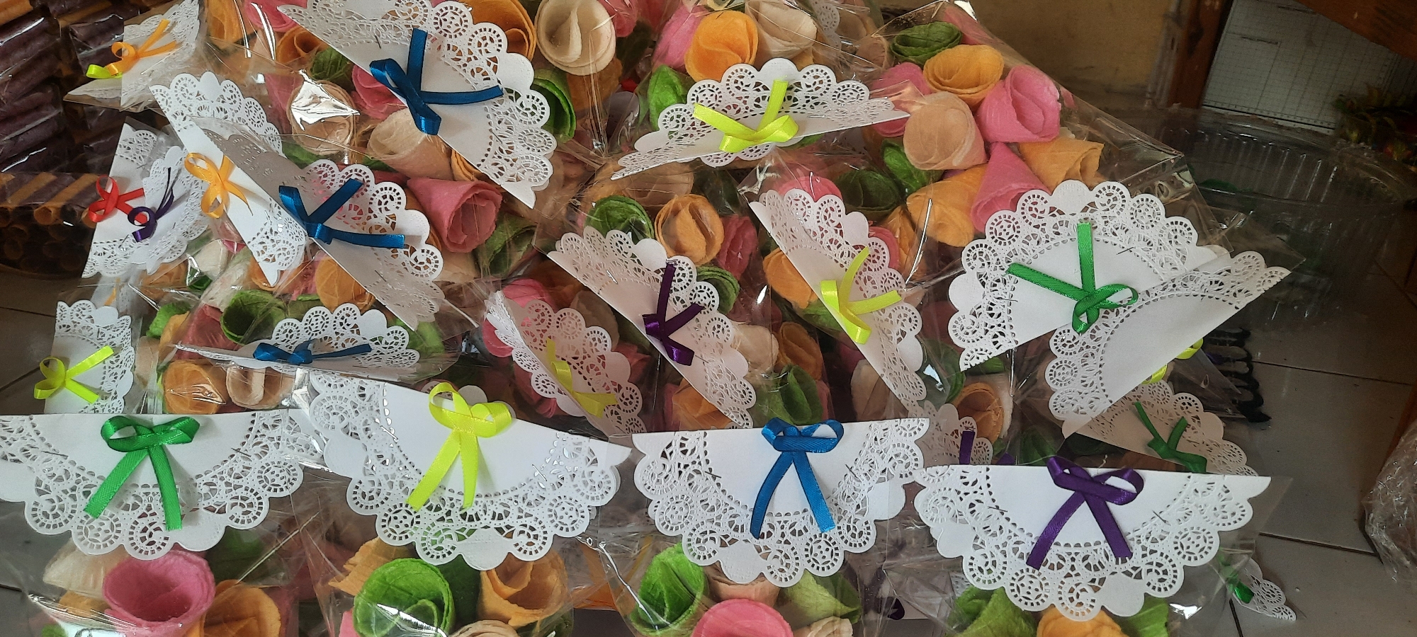 A pile of colorful, cone-shaped snacks are displayed in clear plastic packaging. Each snack is adorned with a white paper doily and a colorful ribbon, creating a festive presentation. The snacks appear to be a variety of pastel colors, including pink, orange, yellow, and green, and are rolled into tight cones. The packaging is arranged in a layered, overflowing manner, suggesting a large quantity. In the background, some brown, rolled items are visible in what appears to be a basket or container. The scene is likely a display of food items for sale, possibly in a market or shop. This particular image is sourced from Ploso, Indonesia.