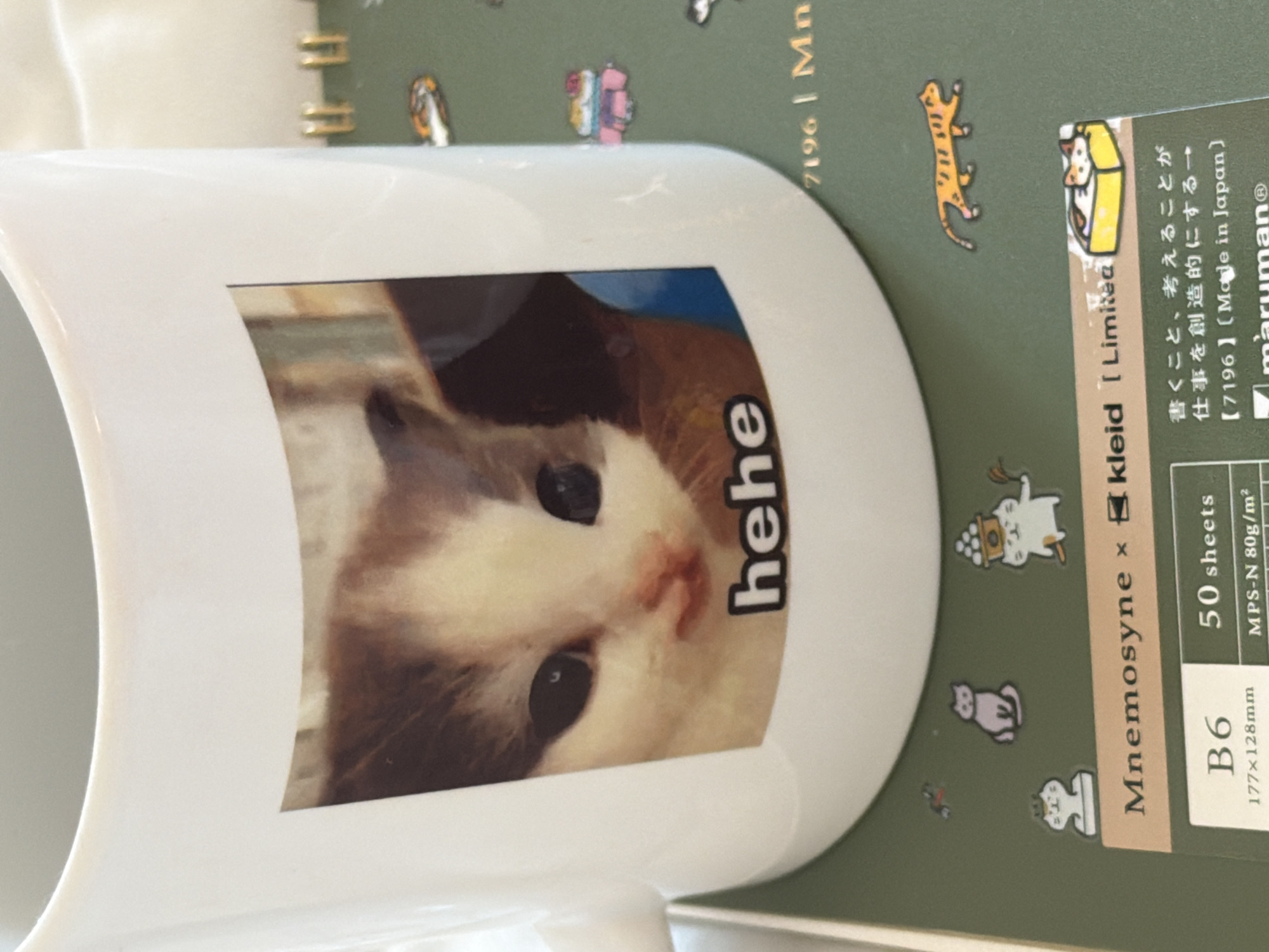 A close-up, top-down shot captures a white mug resting on a green, spiral-bound notebook. The mug features a printed image of a light-colored cat with brown patches and dark eyes, gazing forward. The word "hehe" is printed in black, block letters next to the cat's face.
The notebook, partially visible, has a dark green cover and gold spiral binding. It is adorned with several small, cartoon-style cat illustrations, including a striped cat, a cat in a box, a cat wearing a chef's hat, and a cat holding grapes.
Visible text on the notebook includes "Mnemosyne x kleid [Limited", "B6", "50 sheets", "177x128mm", "MPS-N 80g/m²", and "maruman®". Japanese text is also present: "書くこと、考えることが 仕事を創造的にする→" (meaning "Writing and thinking make work creative →") and "【7196】(Made in Japan)". A partial text "7196 | Mn" is also visible on the green background. The background is light-colored and out of focus, suggesting an indoor setting like a desk or tabletop.