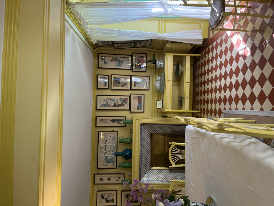 The interior of a room in a house in Giverny, France, located at latitude 49.076 and longitude 1.534. The room is painted yellow, and the walls are adorned with numerous framed prints, primarily of Japanese art depicting birds and nature scenes. A fireplace with a tiled surround sits to the left, and a wooden table with two shelves sits to the right. A white porcelain cat sits on the top shelf of the table. The room has a checkered tile floor in shades of red and cream, and a large white tablecloth sits on a table with four yellow chairs. There is a single white chair with a straw seat on the far right of the room. A window with white lace curtains provides light to the room, and a potted plant with pink flowers sits on the table. There is a small table with a white vase on top, and the room is otherwise empty.