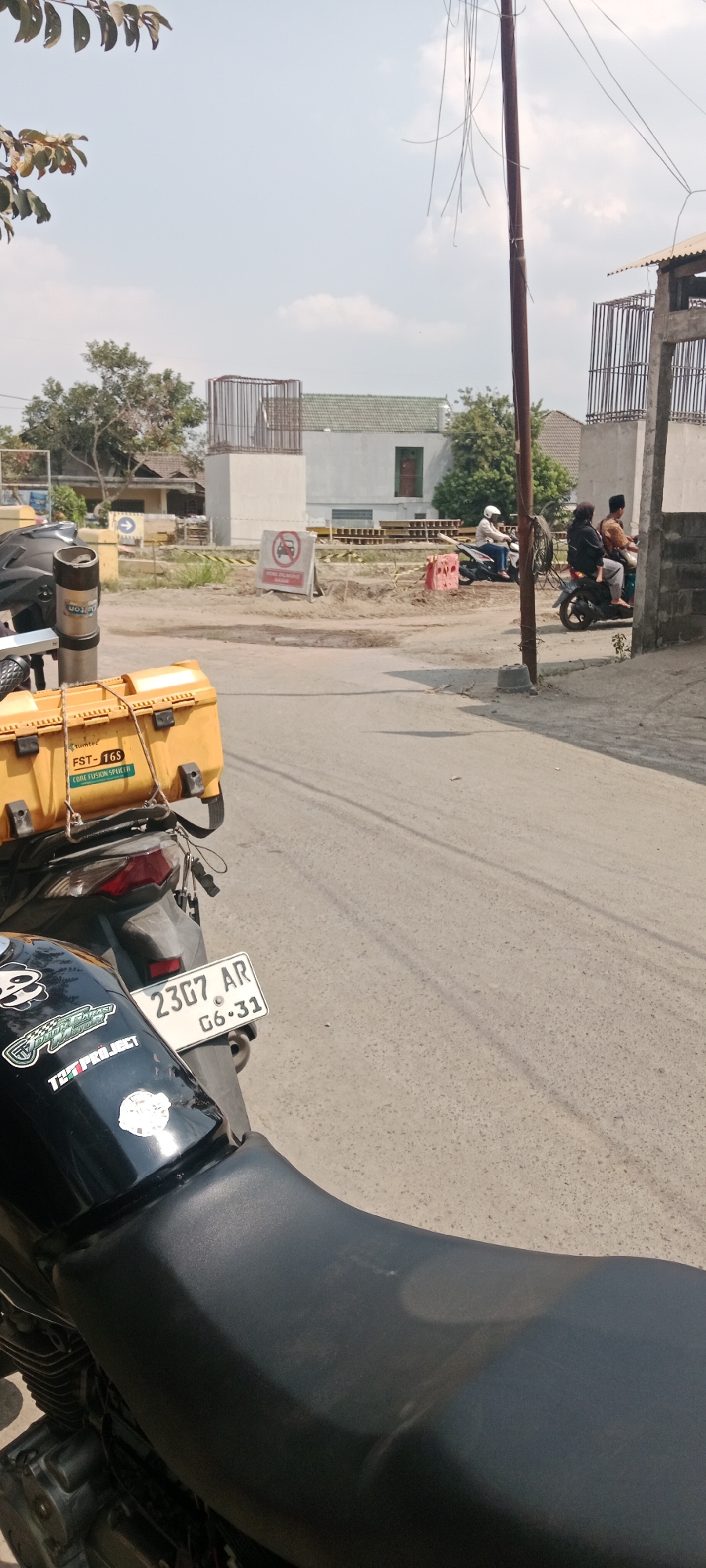 In Purwomartani, Indonesia, a motorcycle is parked on the side of a road undergoing construction. The motorcycle has a yellow storage box mounted on the back, a license plate that reads "2307 AR G6.31", and several stickers on its fuel tank, including a panda sticker. In the background, there are construction materials, a sign prohibiting motorcycles, and two motorcycles with riders. The sky is clear with some clouds, and a utility pole with dangling wires stands prominently in the scene.