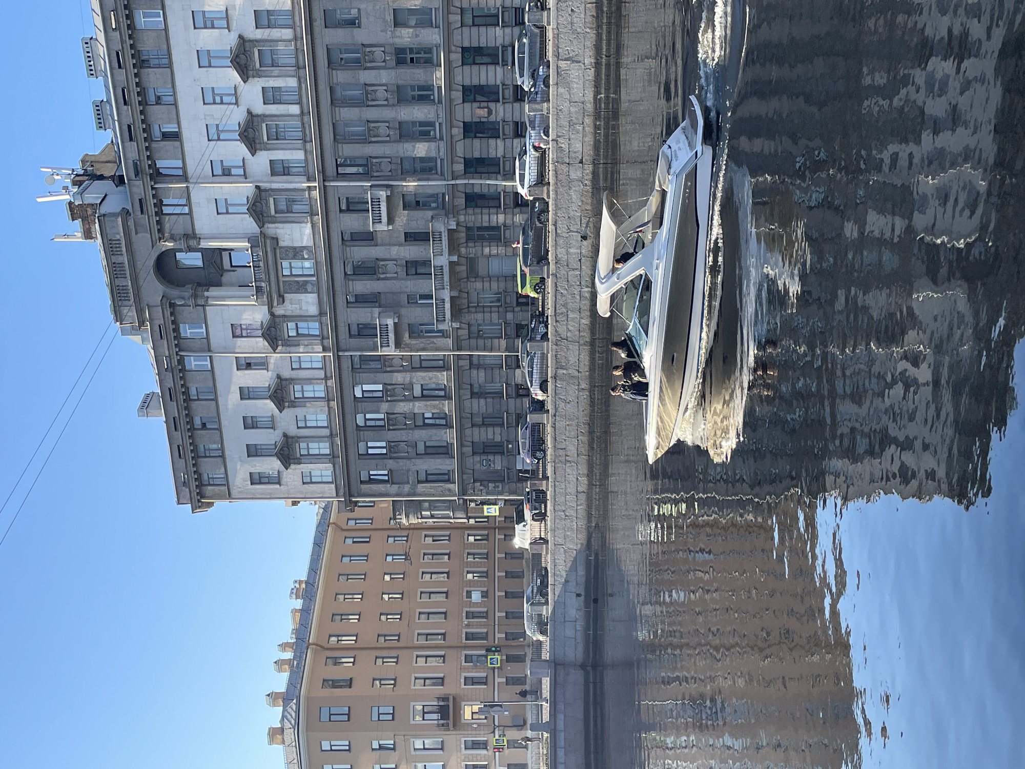 The image depicts an urban scene along the Griboyedov Canal Embankment in Saint Petersburg, Russia, under a clear blue sky. A modern motor yacht, predominantly white with a tan hull stripe and a white canvas bimini top, is moving rightward across the canal's foreground, leaving a distinct wake. Approximately 4-5 individuals are visible seated within the yacht's cockpit.
On the immediate right bank of the canal, a large, multi-story building with a grey, classical facade features an arched portico flanked by columns on an upper level, along with numerous windows and decorative details. Its roof is equipped with several communication antennas. Opposite, on the far bank, a multi-story building with a lighter, beige-colored facade is visible.
A granite embankment wall separates the canal water from a paved roadway. Along this road, multiple vehicles, including sedans and SUVs, are either parked or in transit. Pedestrian crossings, traffic lights, and street furniture are visible. A few indistinct figures are present on the pedestrian walkways adjacent to the road. The sunlight casts reflections of the buildings onto the canal's surface.