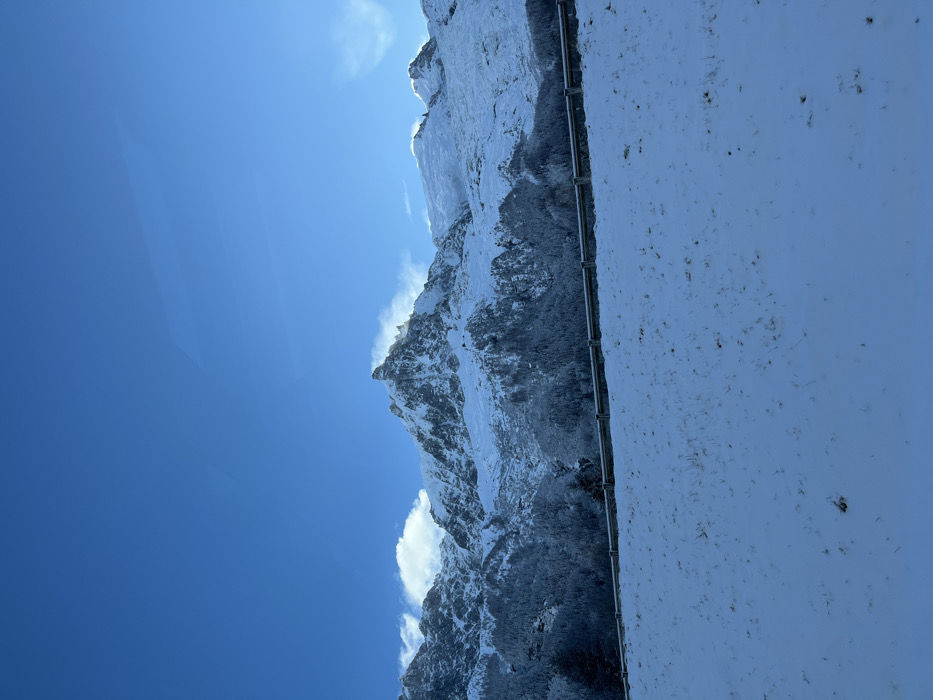This image captures a magnificent winter mountain landscape under a clear blue sky, set in Les Houches, France.
The scene is dominated by towering, rugged mountains extensively covered in brilliant white snow and ice. These snow-capped peaks stretch across the middle and right side of the frame. Darker, rocky sections and sparse conifer trees, also dusted with snow, are visible on the slopes, providing contrast to the pristine white. Wisps of cloud or wind-blown snow cling to some of the higher ridges, adding to the dynamic feel of the high-altitude environment.
A vast, deep blue sky fills a significant portion of the upper left, indicating a bright, sunny day. The sunlight strongly illuminates the snow, creating a crisp and cold atmosphere. A few thin, white clouds are scattered in the sky, particularly near the mountain tops.
In the foreground, a flat, snow-covered surface occupies the right third of the image. A dark, thin metal railing or barrier, possibly part of a viewing platform, balcony, or mountain road, runs horizontally across this snow-covered ground. The snow on this surface is speckled with small dark specks, likely dirt or debris.
No people or explicit activities are visible, suggesting a serene moment of observation of the natural grandeur. The image evokes feelings of awe, tranquility, and the pristine beauty of an alpine winter. The clear conditions suggest mid-morning or early afternoon. There is no visible text in the image.