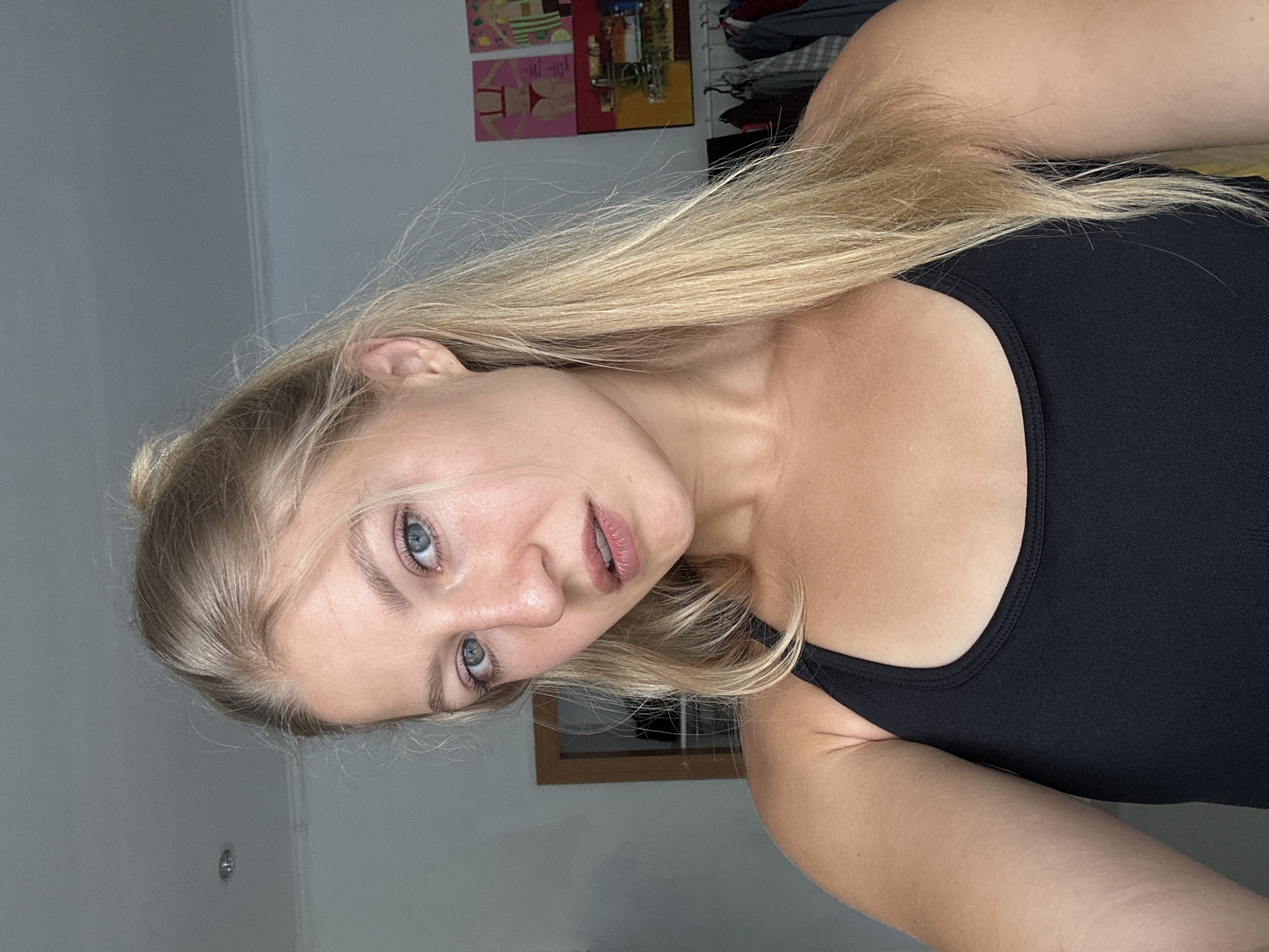 A young woman is captured in a close-up, chest-up shot, her head tilted, looking directly into the camera with a soft, neutral expression. She has long, straight blonde hair cascading over her shoulders and chest. Her eyes are a striking blue, and her lips have a natural, light pink hue. She is wearing a plain black tank top or camisole.
The image is oriented horizontally, suggesting it might be a selfie taken with the phone tilted or while lying down. The background behind her consists primarily of a light gray wall. In the upper right portion of the frame, a partially visible clothes rack holds various garments. Above the rack, two framed art pieces are displayed; one features abstract shapes in reds and yellows, while the other shows more figurative elements, including what appears to be a bikini top and a cocktail glass. A small, rectangular wooden-framed object, possibly a mirror or a window, is visible in the lower central background, reflecting a grid pattern.
The lighting is soft and even, casting minimal shadows and making the exact time of day indeterminate, though it appears well-lit. The scene depicts an indoor setting, likely a room within a residence in Kuta Selatan, Indonesia. There is no visible text in the image.