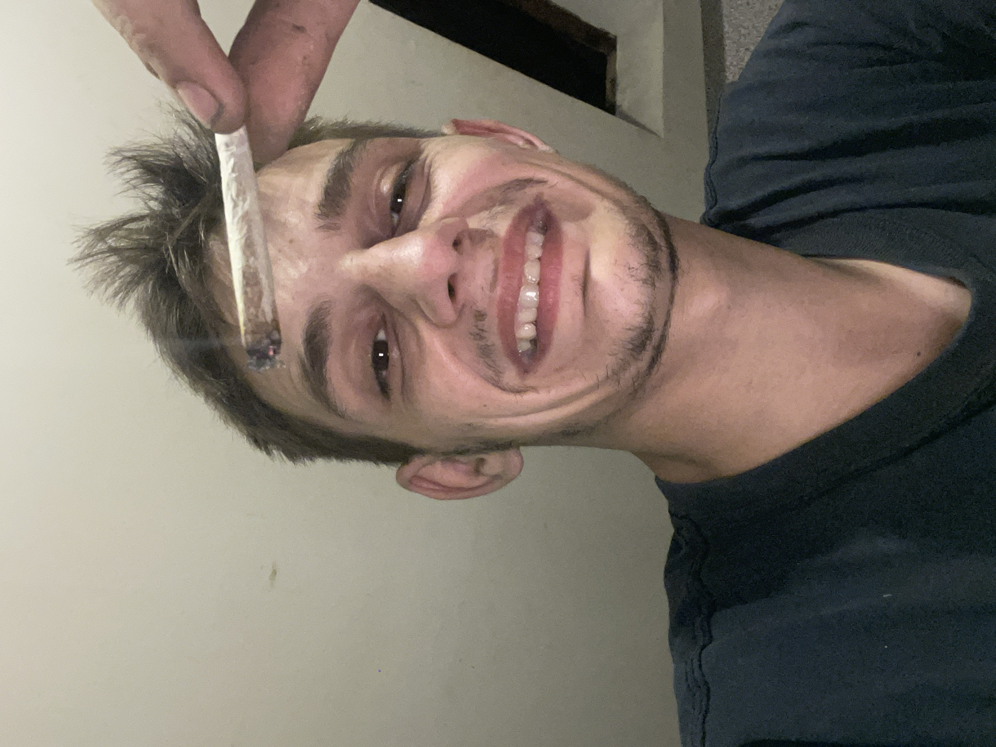 In a close-up, low-angle shot, a person with short, messy brown hair and a light beard smiles broadly, holding a lit cigarette between their thumb and forefinger just above their forehead. The person is wearing a dark t-shirt. The background is a plain, textured wall, likely indoors, with a hint of a dark, angled structure visible in the upper right corner. The lighting is dim, with a warm tone, suggesting an indoor environment. The image appears to be a selfie.