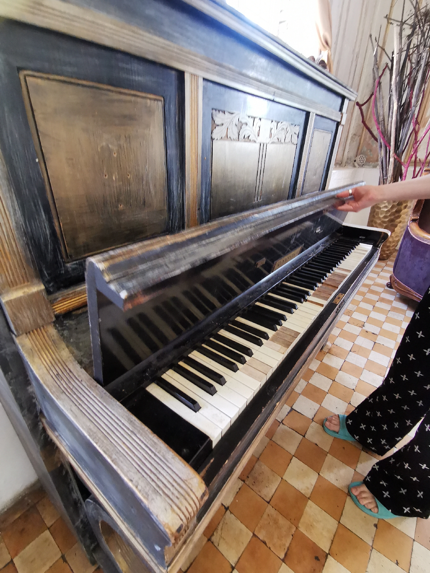 The image shows an upright piano in what appears to be a room with terracotta-colored checkered tile flooring. The piano is aged and has some missing or damaged ivory keys. The case is painted in dark blue with gold decorative accents. A person, visible from the mid-arm down, is opening the piano's fallboard. The person is wearing dark pants with a light-colored cross pattern and light blue open-toed sandals. In the background, there is a decorative golden vase with branches, and a portion of a purple chair is visible. The specific location within Moscow, Russia, is not discernible from the provided image.