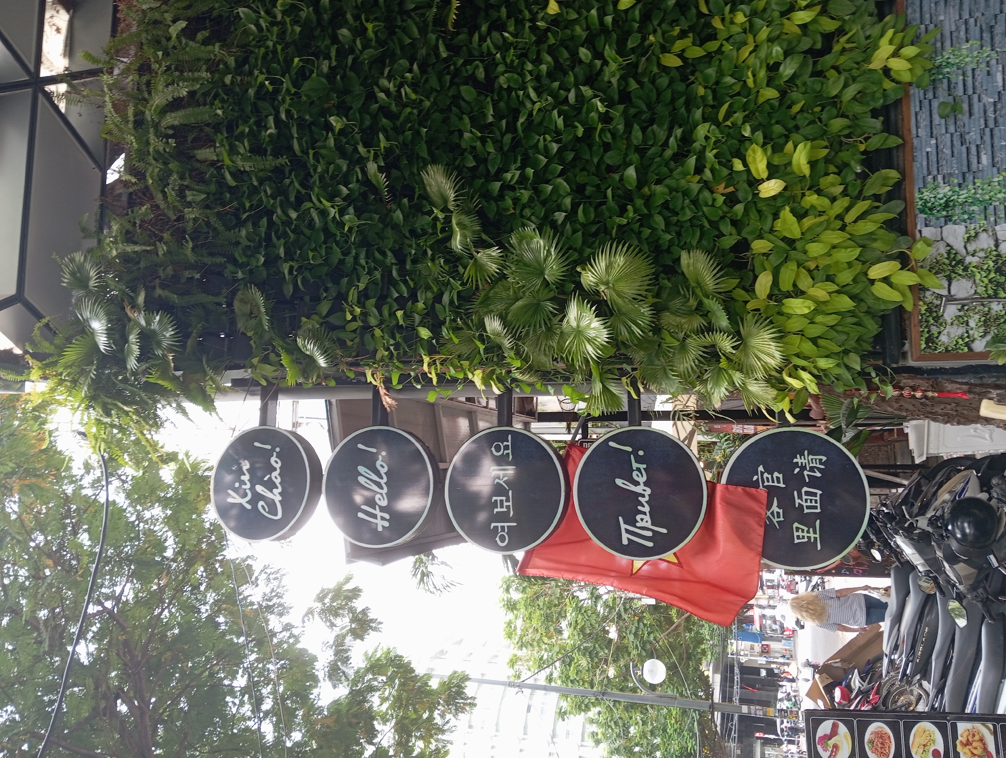 The image depicts a street scene in Nha Trang, Vietnam, characterized by lush greenery and a collection of circular signs indicating greetings in multiple languages. The signs are stacked vertically and feature text in Vietnamese ("Xin Chào!"), English ("Hello!"), Korean ("여보세요"), Russian ("Привет!"), and Chinese ("客官里面请"). Adjacent to the signs, a Vietnamese flag is prominently displayed. In the background, a woman with blonde hair is walking away from the camera, and various motorcycles are parked. The environment includes trees, buildings, and a vertical garden feature with diverse plant life.