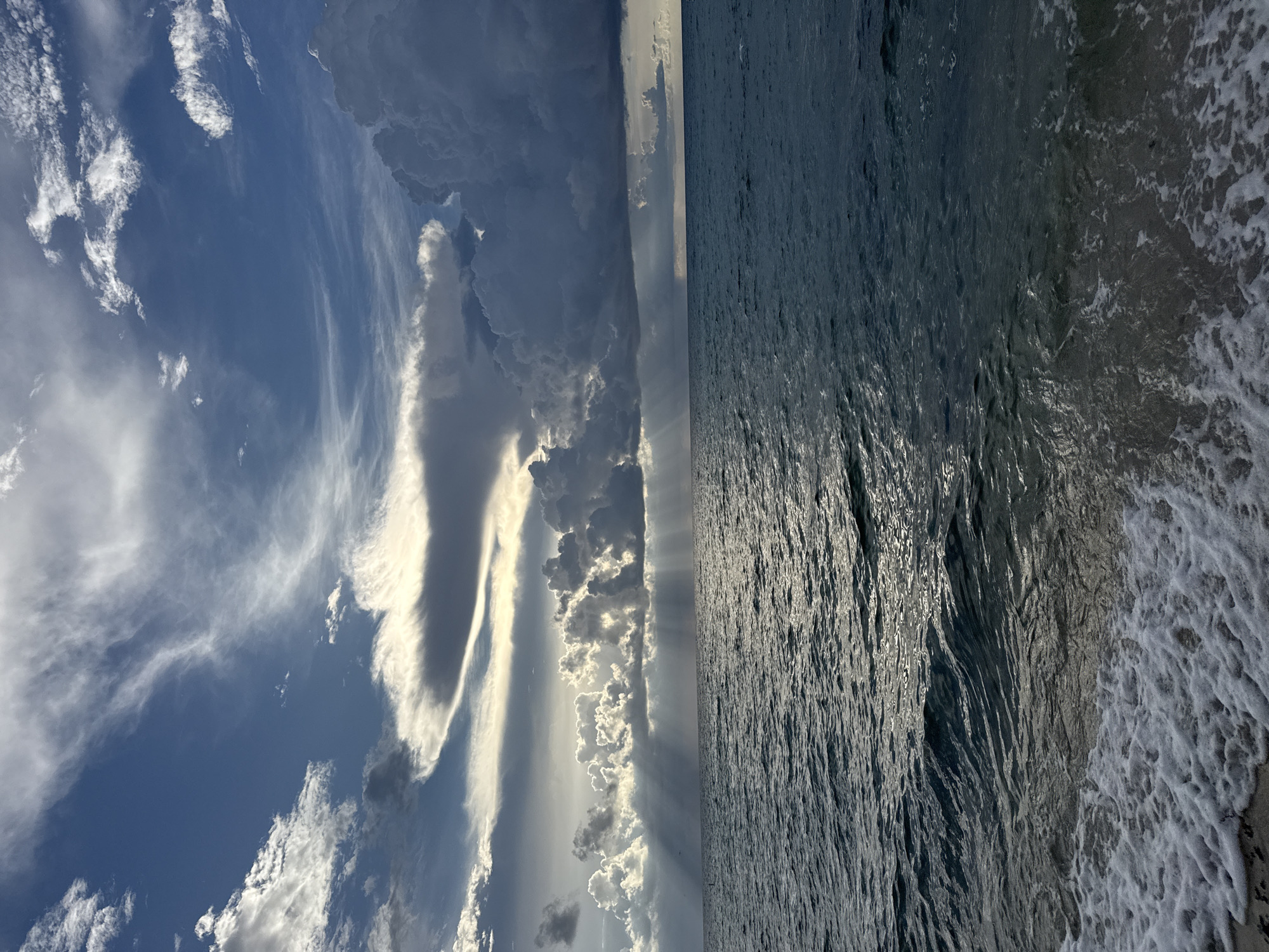 This is a photograph of the ocean and sky at Sunny Isles Beach, Florida, United States of America. The scene is dominated by dramatic clouds and a powerful juxtaposition of light and shadow over the water.
The sky is a complex tapestry of clouds. In the upper portion of the frame, wispy, cirrus clouds stretch across a bright blue sky, tinged with sunlight. Further down, larger, more textured clouds appear, some dark and foreboding, others illuminated from behind by the sun, creating a dramatic, almost theatrical effect. Rays of sunlight pierce through breaks in the clouds, casting beams onto the ocean below. The clouds on the right side of the image are particularly dark and voluminous, suggesting an approaching storm or the aftermath of one.
The ocean occupies the lower half of the image. The water is a deep, moody blue-green, with visible ripples and waves. Sunlight reflects off the water's surface, creating streaks of shimmer and highlighting the texture of the waves. In the foreground, a small section of beach is visible, with white foam from the breaking waves washing ashore. The sand appears dark and wet.
There are no people or man-made structures clearly visible in the scene, focusing the viewer's attention entirely on the natural elements of the sky and sea. The overall impression is one of natural grandeur and the dynamic interplay of weather phenomena over the ocean.