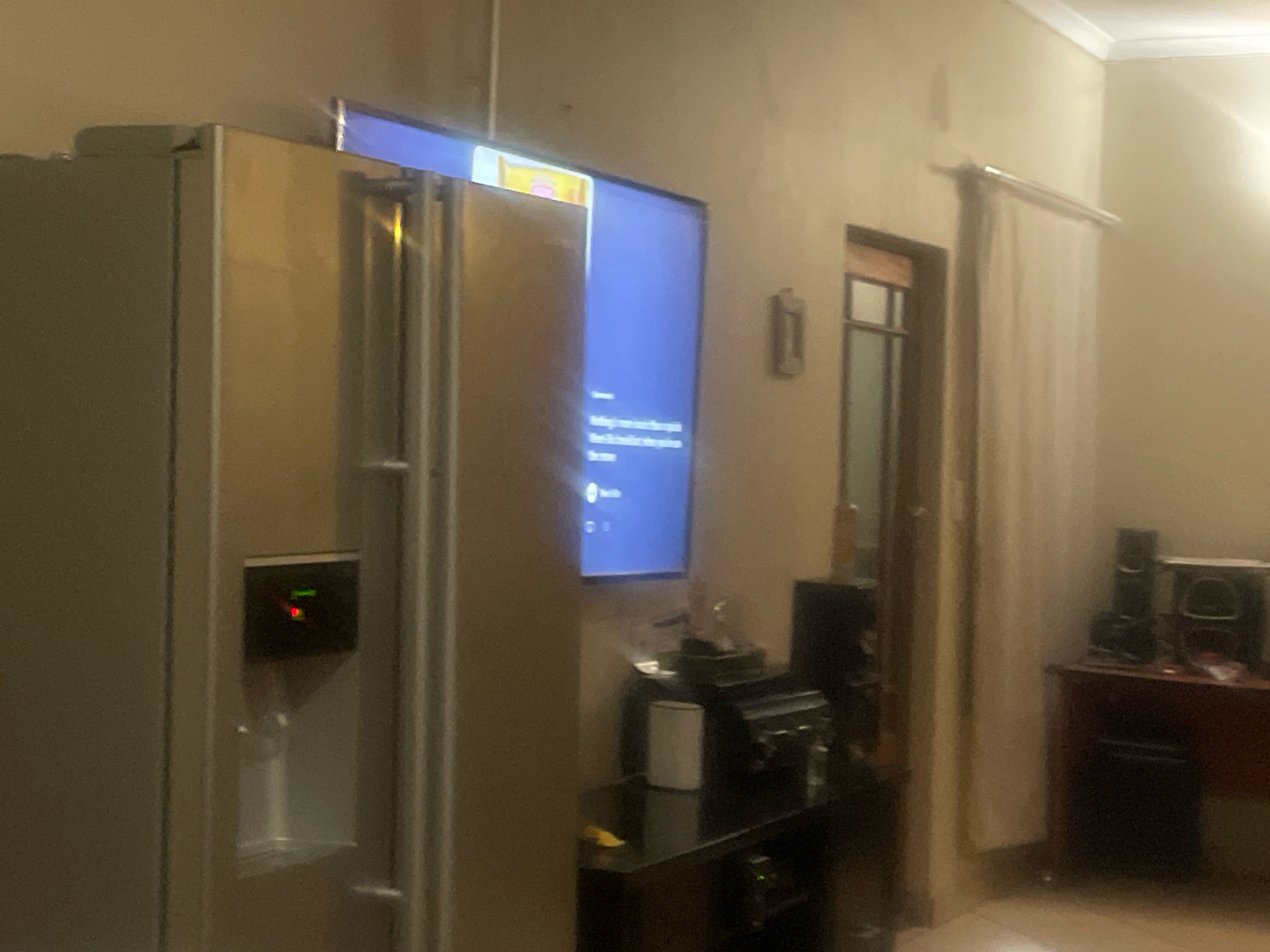 In a dimly lit room, a large stainless steel refrigerator stands prominently on the left side of the frame. A television screen is mounted above the refrigerator, displaying a blue interface with text. To the right of the refrigerator, a black entertainment stand holds various electronic devices, including what appears to be a printer and speakers. Further to the right, a dark wooden table or desk is visible with more electronic equipment, including speakers. A light-colored curtain hangs on the right side of the room, partially obscuring a window. The walls are painted a neutral, light color. The scene appears to be an interior space, possibly a living room or kitchen area, in Bronkhorstspruit, South Africa.