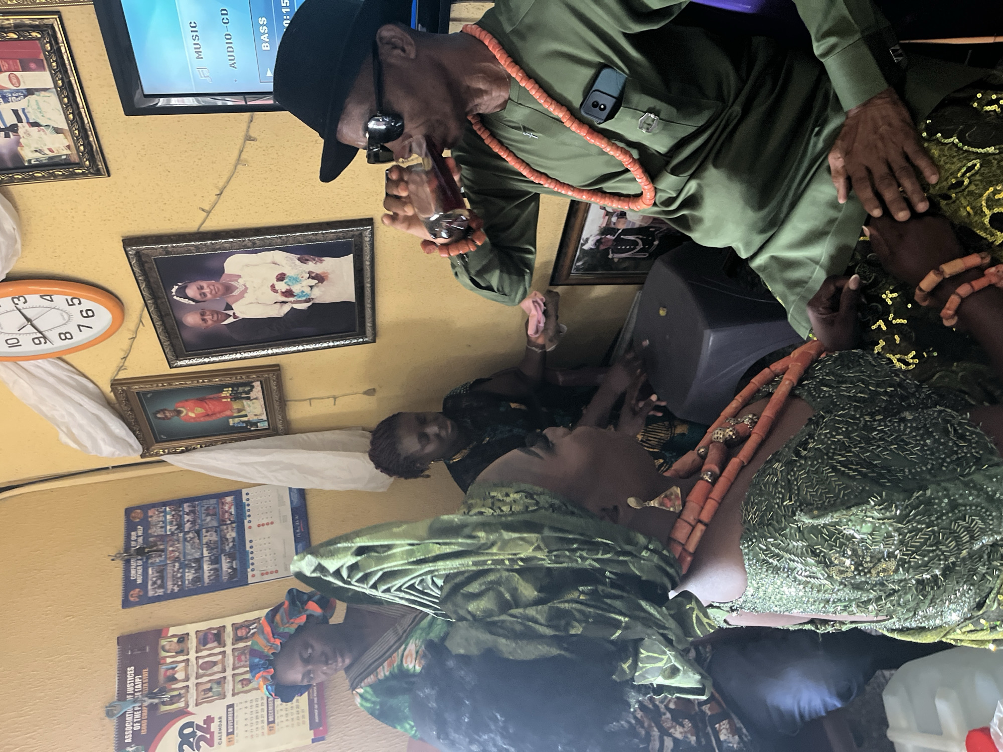 The image depicts an interior scene in what appears to be a residential setting in Warri, Nigeria. An older man in a green shirt, black hat, and coral beaded necklace is drinking from a glass while standing near a seated woman adorned in green traditional clothing and coral beads. A second woman sits behind the first. The room has yellow walls decorated with framed pictures, a clock, and calendars. A television is playing audio in the background. There is interaction between the man and the seated woman, with the man’s hand near her head.