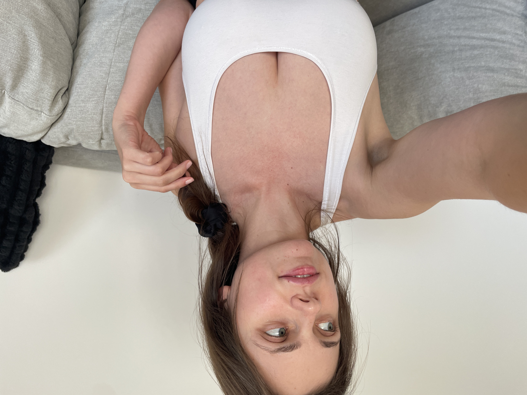 A young woman with brown hair braided into a ponytail, secured with a black scrunchie, is seen from the chest up. She is wearing a white sports bra and is seated on a grey couch. Her left arm is extended towards the viewer, as if holding a camera. She is looking to her left with a slight smile. The background consists of a plain white wall and a portion of a dark, textured blanket on the right side of the frame. The scene appears to be indoors, in a domestic setting.