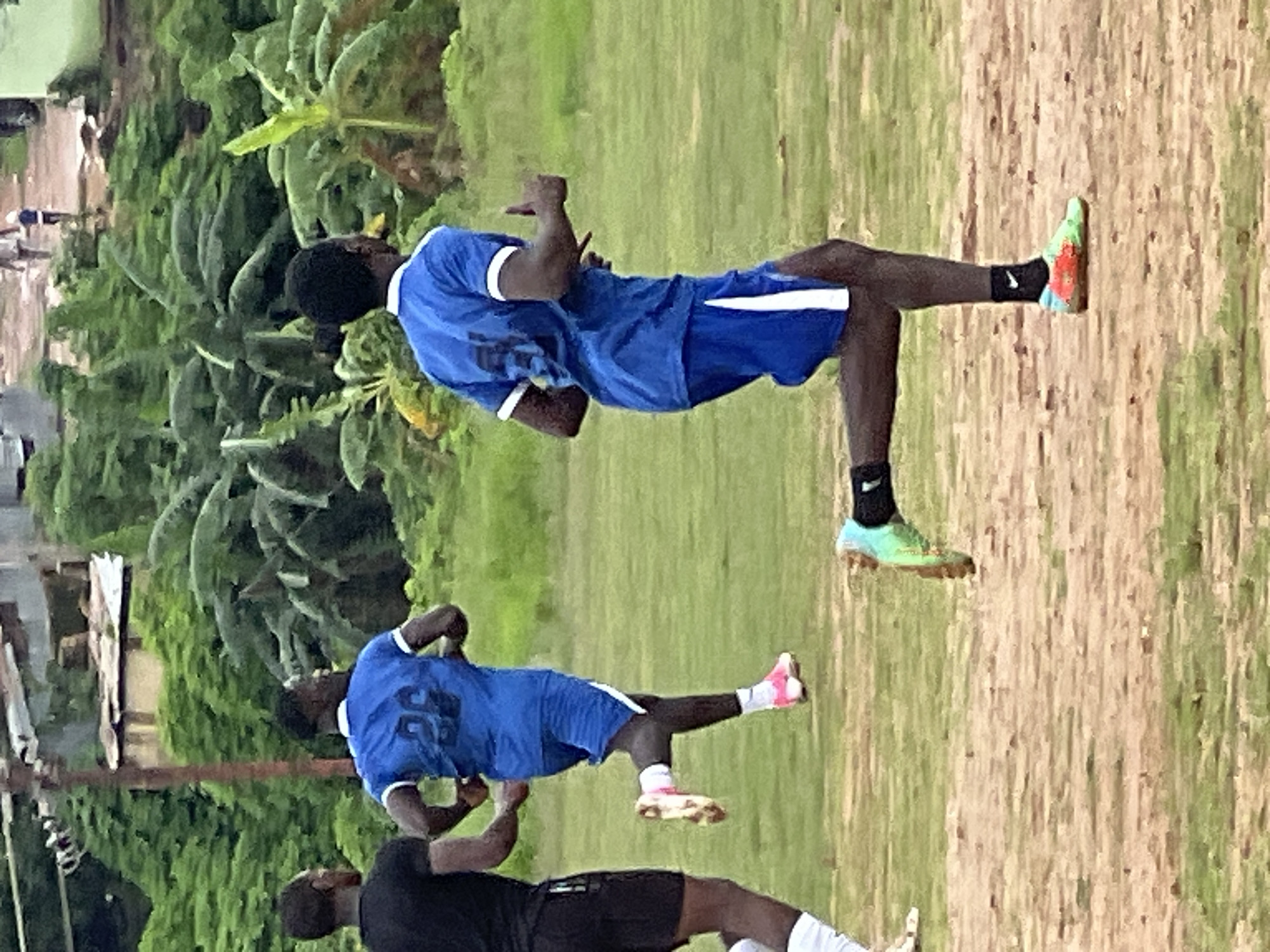 In an outdoor scene, three young Black men are running on a grassy field, possibly engaged in athletic training. The man in the foreground, wearing a blue jersey with white trim and the number "29" on the back, along with blue shorts with white stripes, is captured in mid-stride. His right leg is extended forward, and his left leg is bent, with a green and red athletic shoe on his foot. He is looking to his right. To his left and slightly behind him, another man in a blue jersey with white trim and blue shorts with white stripes, also numbered "29", runs with his left leg forward and right leg bent. He is looking to his left. Further to the left and behind these two, a third man, wearing a black t-shirt and black shorts, runs with his left leg forward and right leg bent. His head is turned to his right, looking towards the other runners. The environment is characterized by lush green vegetation and trees in the background, with a dirt and grass field in the foreground. A dirt path with some scattered debris is visible in the far background, along with structures that suggest an urban or semi-urban setting. The sky is overcast. This scene is likely located in Jos, Nigeria, given the context of the request, suggesting a common practice of outdoor sports or training in such environments within the city.