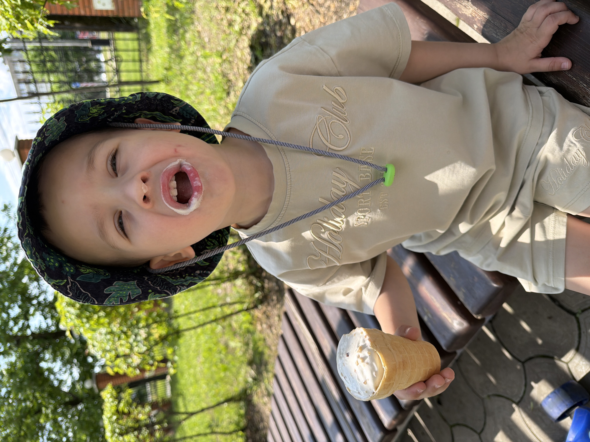 A young boy wearing a bucket hat and a light beige t-shirt and shorts is sitting on a wooden bench outdoors, holding an ice cream cone. He has ice cream on his face around his mouth. The background consists of green trees, a fence, and a building. The ground is paved with hexagonal tiles. It is a sunny day. The image was taken in Khabarovsk, Russia.