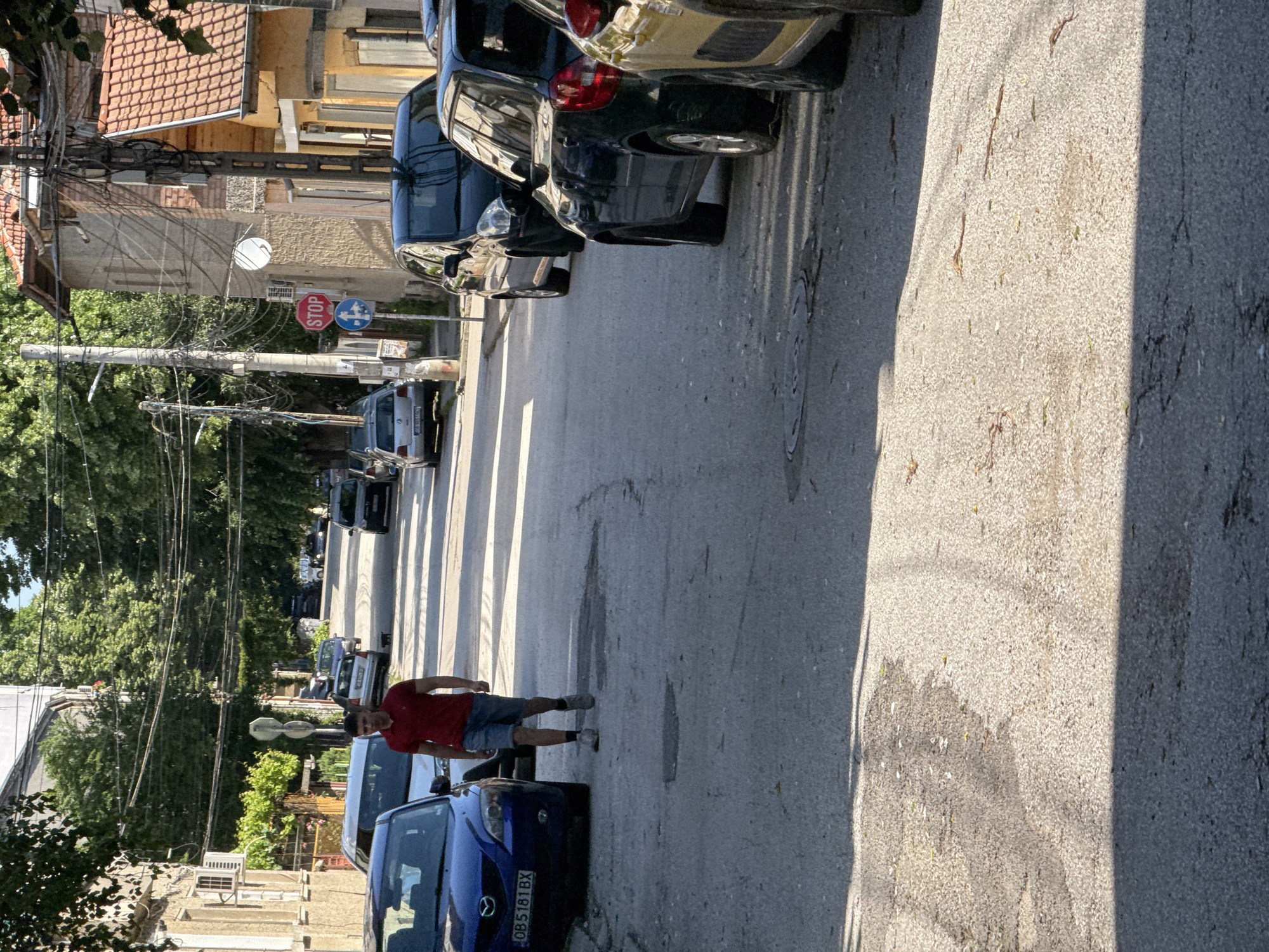 The image depicts a residential street scene in Lovech, Bulgaria. A man in a red shirt and shorts is walking across a pedestrian crosswalk in the street. Various parked cars line both sides of the road. A blue Mazda with license plate OB5181BX is visible on the right side of the road closest to the crosswalk. Utility poles with numerous wires run along the side of the road on the left side of the frame. A stop sign is partially visible in the background. Buildings with tiled roofs are present on either side of the street. The street surface is asphalt, showing signs of wear and repair. The weather appears sunny, casting shadows on the road.