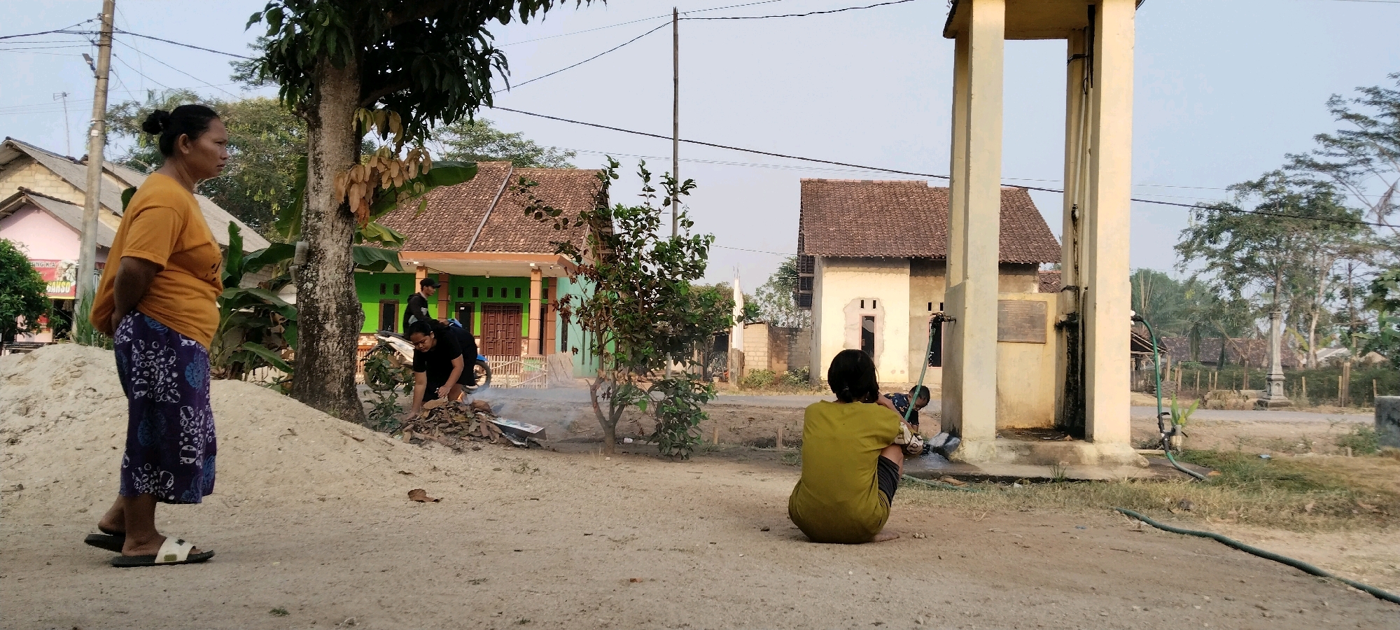 The image depicts a street scene in Tanjung Ratu Ilir, Indonesia. On the left, a woman wearing an orange t-shirt and a patterned skirt stands facing left, with her hands behind her back. In front of her is a large pile of light-colored sand or soil. To her right, two individuals in black clothing are tending to a small fire burning dry leaves. A white motorcycle is parked behind them. Behind the individuals, a green-painted house with a brown tiled roof is visible, bordered by a wooden fence. Further in the background, other houses with similar tiled roofs are scattered among trees. On the right side of the image, a light-colored concrete structure with two large pillars stands, resembling a water tower or pump station, with a green hose attached. A person in a yellow shirt is seated on the ground facing away from the camera, near the base of this structure. The ground is a mixture of dirt and sand, with sparse vegetation. The sky is clear and overcast, suggesting it is daytime.