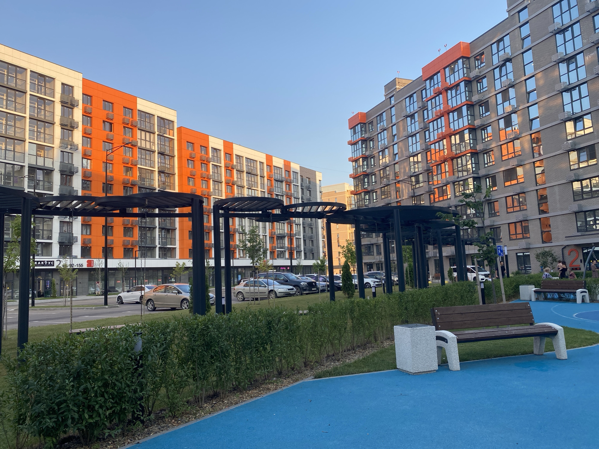 The image depicts a modern residential complex in Krasnodar, Russia, under a clear blue sky, suggesting a daytime setting, likely late afternoon or early evening due to the warm light. The scene features two large apartment buildings; one on the left is predominantly white and orange, while the building on the right is gray with orange accents. Both buildings have numerous windows and balconies, with air conditioning units visible on many.
In the foreground, a blue recreational surface is bordered by a neatly trimmed green hedge and a gravel path. A wooden bench with a concrete trash receptacle is positioned on the blue surface, facing towards the buildings. Another similar bench is visible further in the background.
Behind the hedge, a parking lot is visible with several cars parked, including a silver sedan and a dark SUV. A row of shops with storefronts is located at the base of the left building. One storefront displays text that appears to read "КАРУС" and "2 155".
A few people are visible in the scene, including a person walking near the shops and a group of people sitting on the bench in the background. The overall impression is one of a well-maintained urban living space.