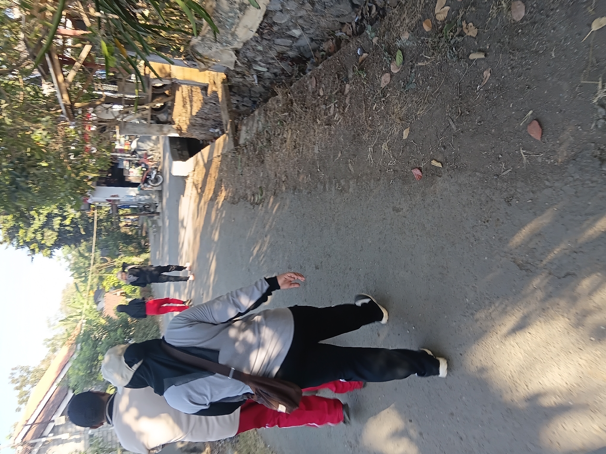 In Tanggul Wetan, Indonesia, four individuals are captured in motion along an asphalt road, their figures silhouetted against the bright daylight. The foreground is dominated by the rear view of a person wearing a light-colored long-sleeved shirt, dark pants, and a baseball cap, with a brown shoulder bag slung across their chest. To their left, another individual in a tan t-shirt and black cap is partially visible. Ahead of them, two more figures are seen walking away: one in a black hijab and red pants, and another in a black hijab and dark pants. The roadside features a narrow, rocky drainage ditch, with sparse vegetation and fallen leaves. In the background, houses with tiled roofs and trees line the street, and a motorcycle is parked near a structure on the right. The overall scene suggests a casual walk in a rural or semi-urban environment.