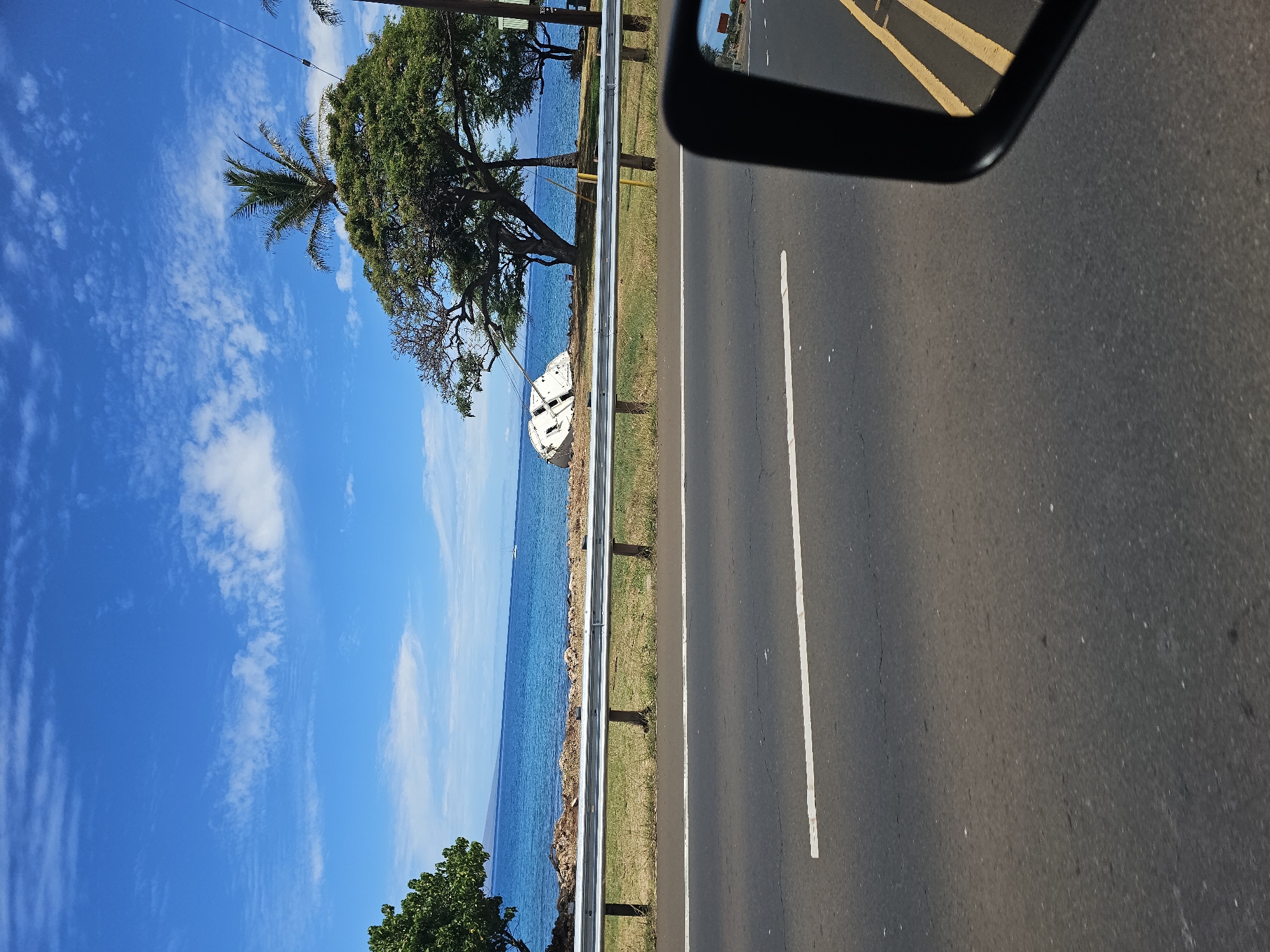 The image depicts a scenic coastal view, likely from a moving vehicle in Lahaina, Hawaii. A bright blue sky with scattered white clouds stretches above a calm, azure ocean. The coastline is lined with green grass and trees, with a prominent shipwreck of a white boat visible on the shore in the mid-ground. A metal guardrail separates the road from the coastal area. The foreground shows a section of asphalt road with a white line marking the lane. A black side mirror of the vehicle is visible in the lower right corner, reflecting a yellow road marking and the ocean beyond. The overall impression is one of a sunny day with a touch of maritime history.